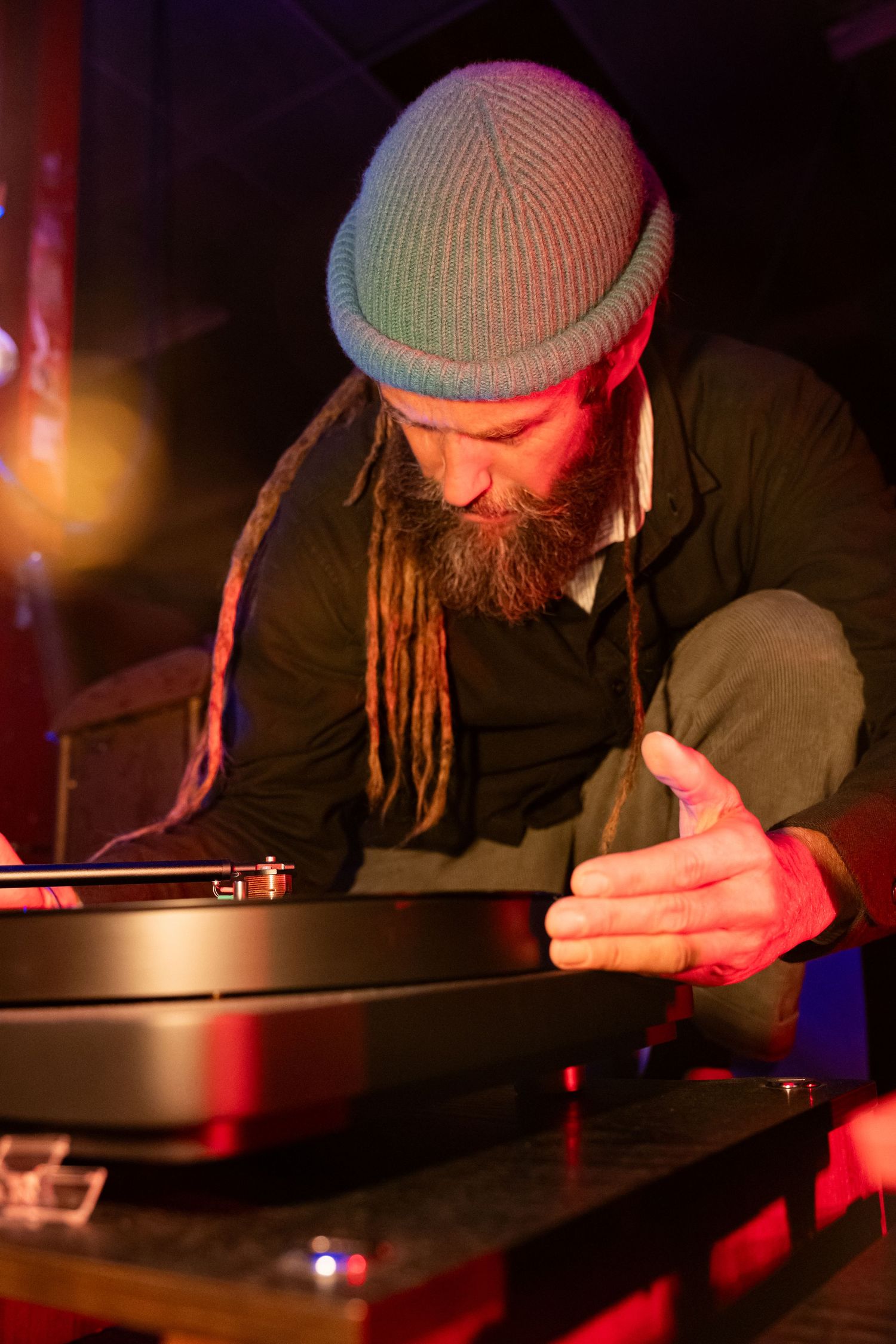 Bearded DJ with dreadlocks and knit hat spinning vinyl records on turntable under red club lighting.