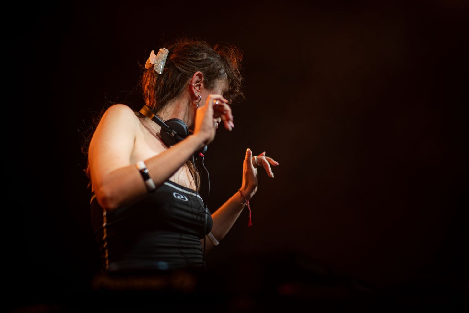 Female DJ performing on dimly lit stage with headphones around neck and hair clip accessory.