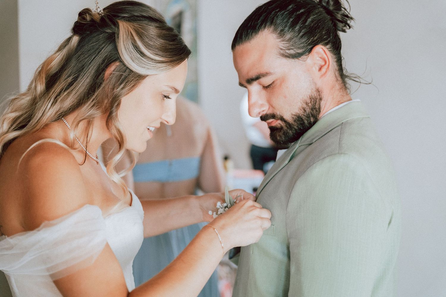 Bride pinning a boutonniere on groom in sage suit during wedding preparations.