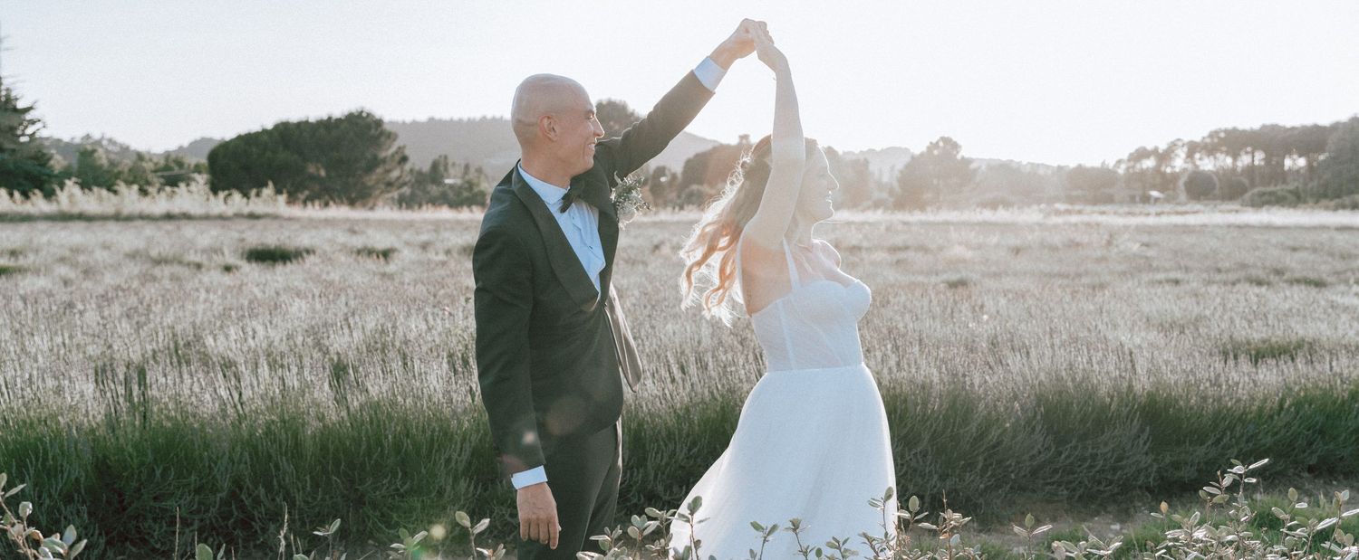 Groom twirling bride in white dress in a sunlit golden field during romantic wedding photo session.