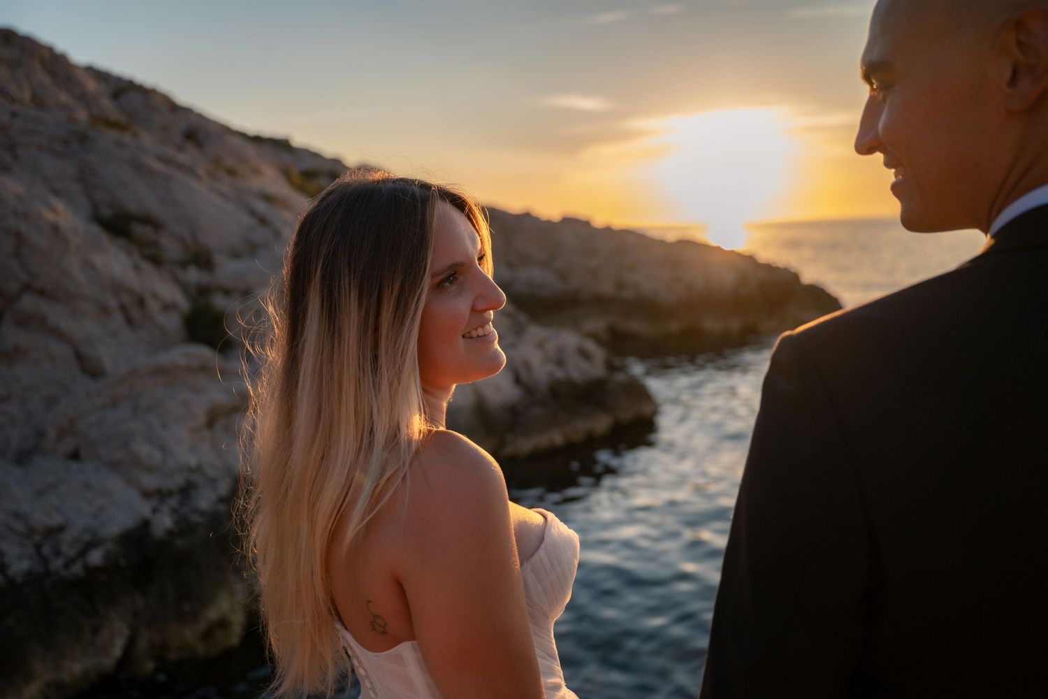 Smiling bride gazing at groom on rocky seaside cliffs at golden sunset.