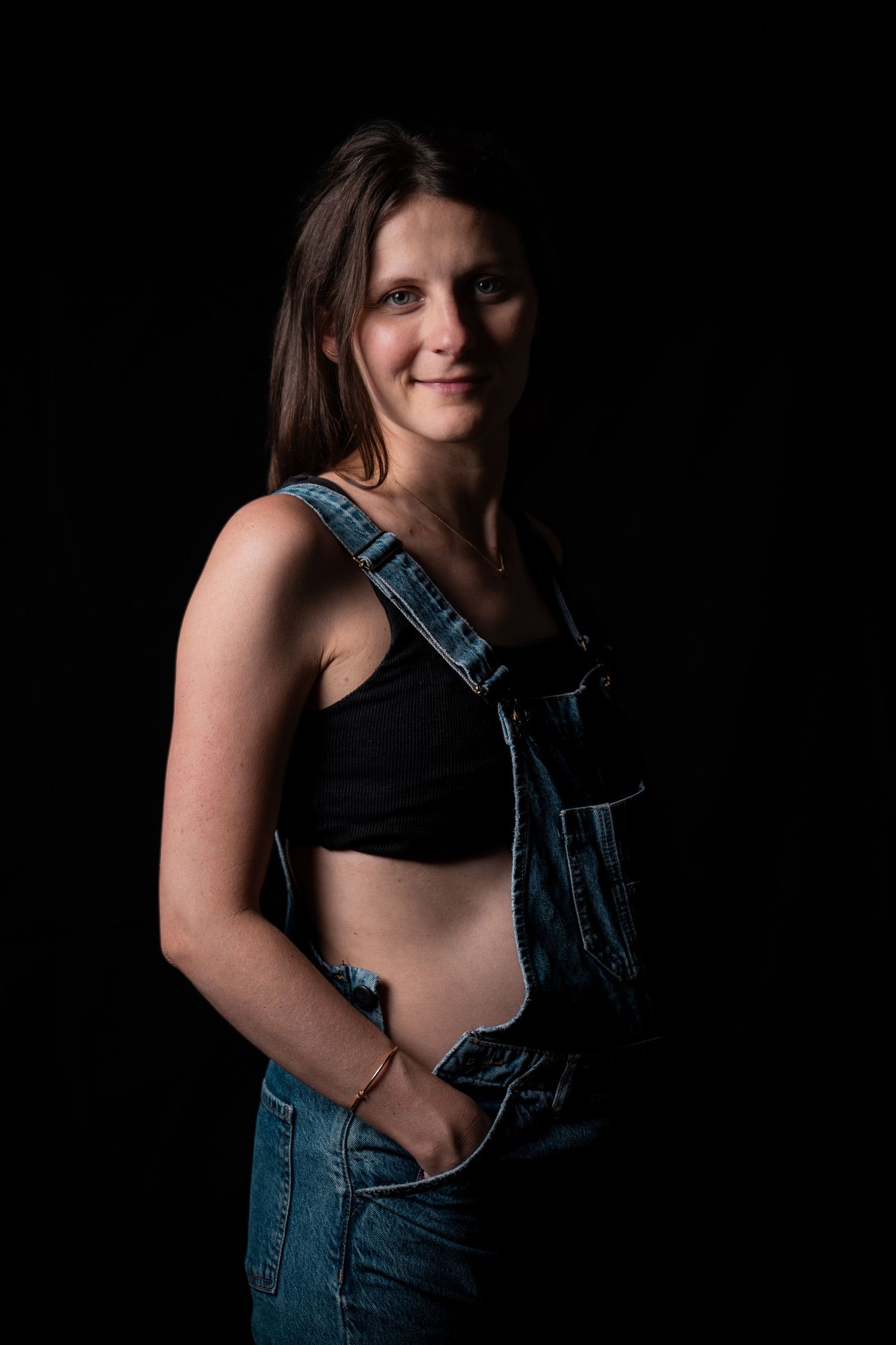 Smiling pregnant woman in denim overalls and black crop top posing confidently against a black background.