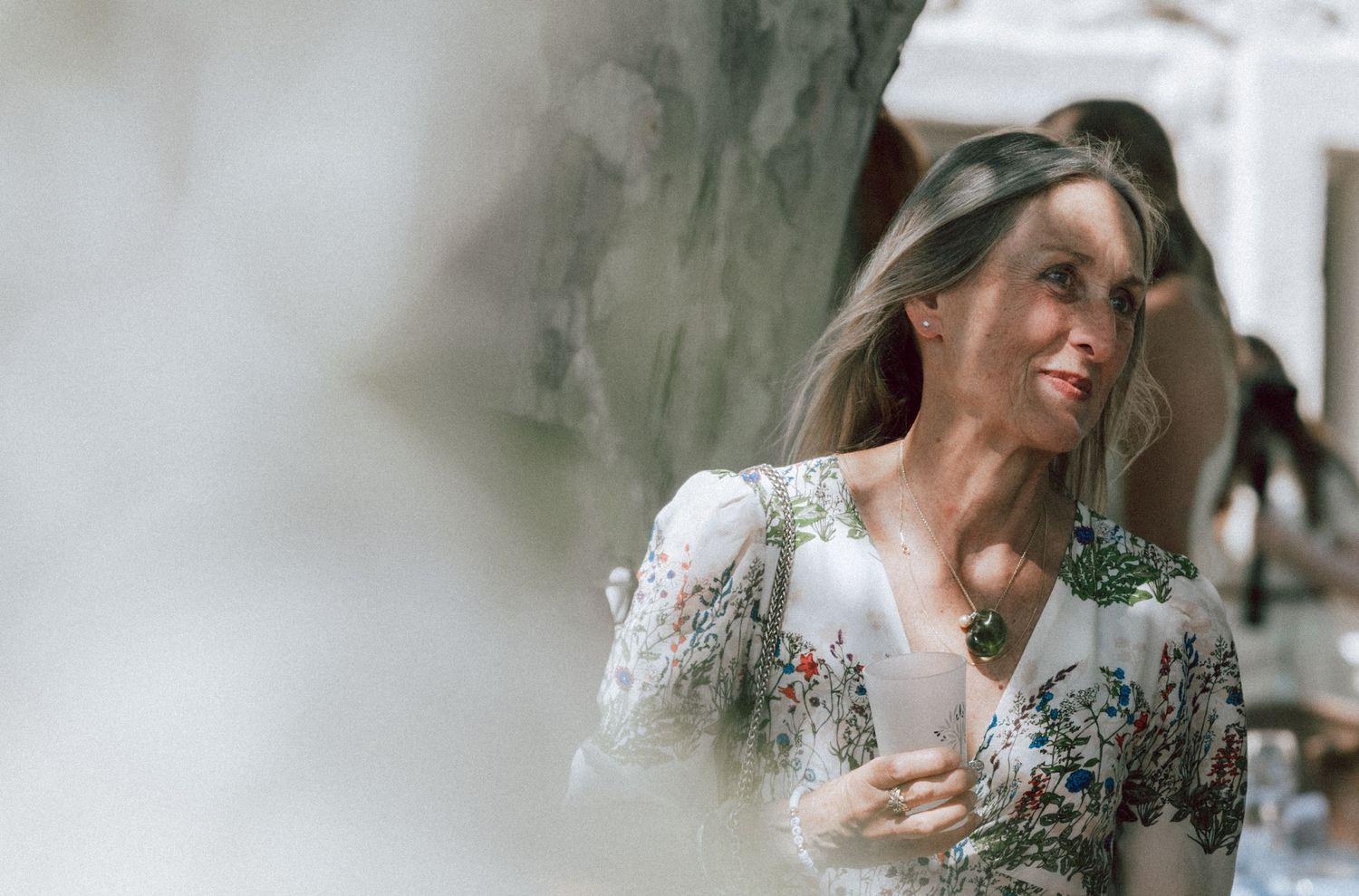 Elegant woman in floral white dress holding a drink and smiling at an outdoor wedding event.