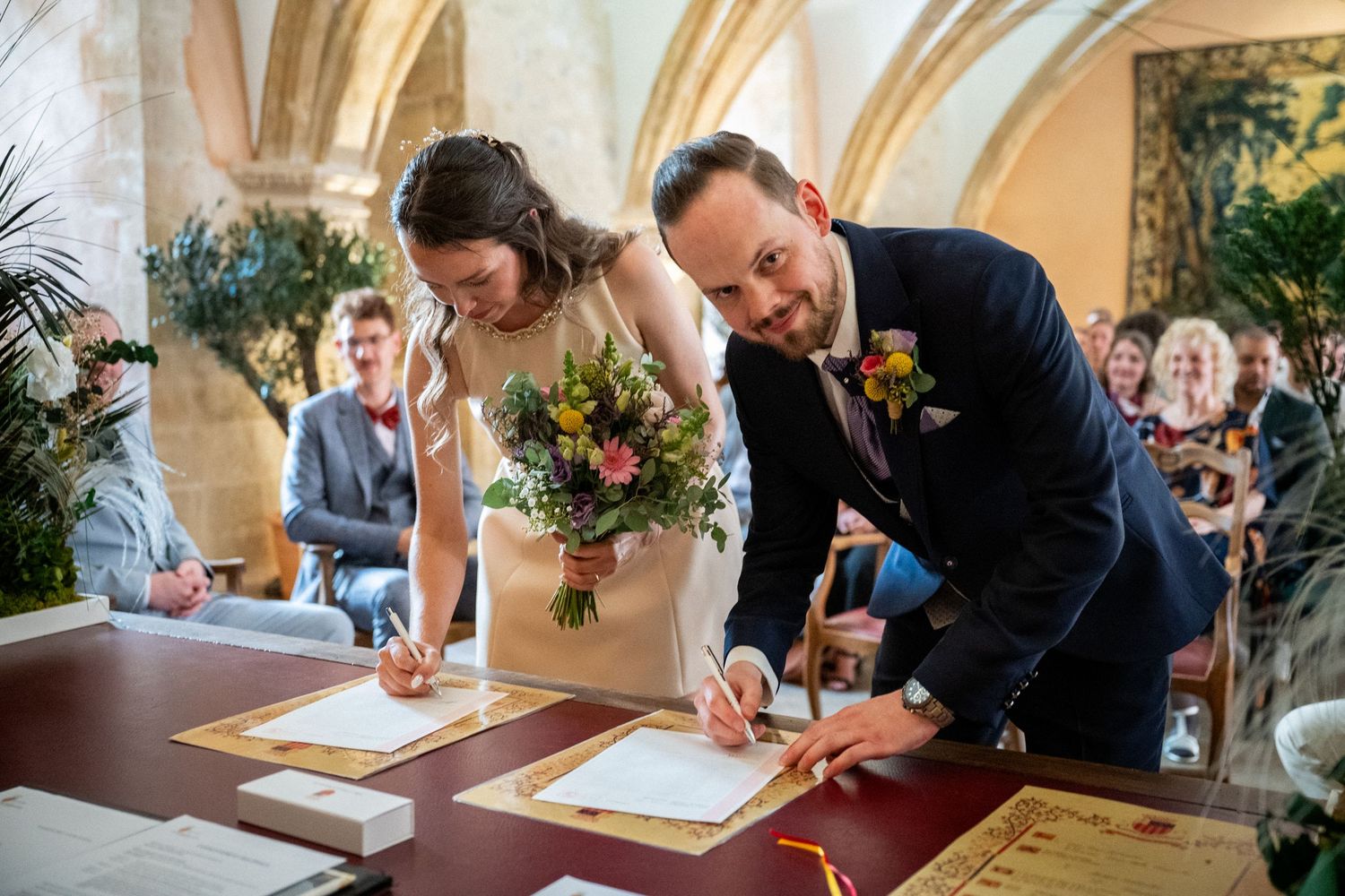 Bride and groom signing wedding documents at ceremony table, bride holding colorful bouquet in elegant venue.
