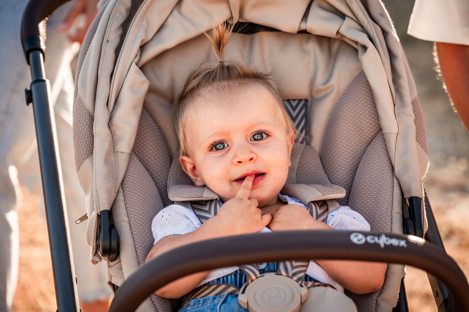 A happy baby sitting in a Cybex stroller, smiling with finger in mouth on a sunny day.