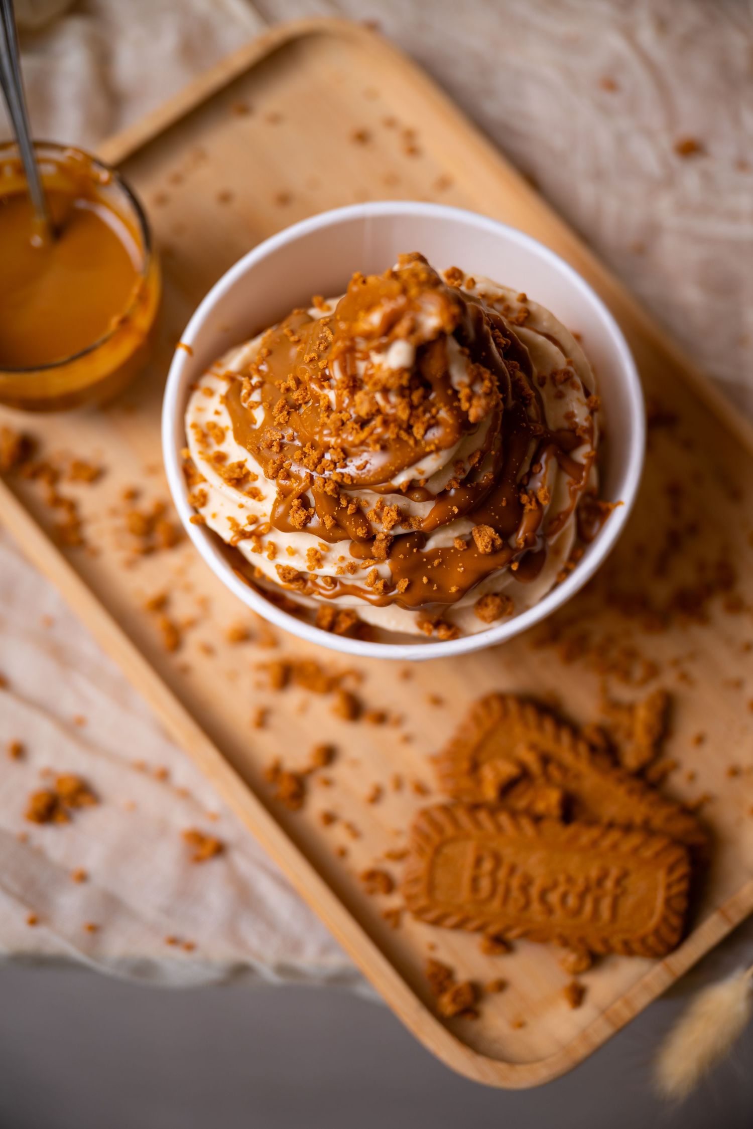 Caramel ice cream topped with crushed Biscoff cookies and caramel sauce in a white bowl on a wooden tray.