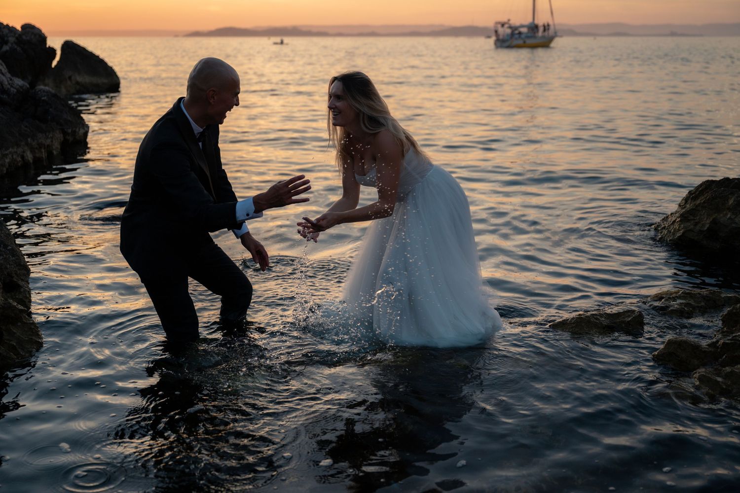 Bride in white gown and groom wade in ocean waves at sunset near rocky shore with sailboat in background.