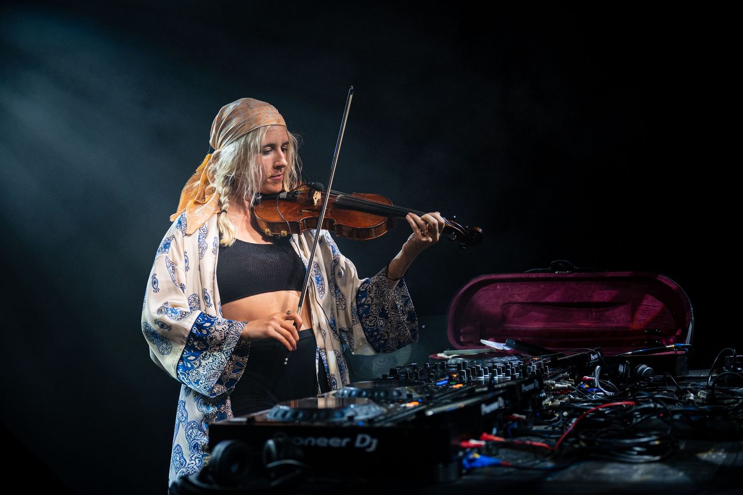 Woman playing violin over Pioneer DJ decks during a smoky stage performance.