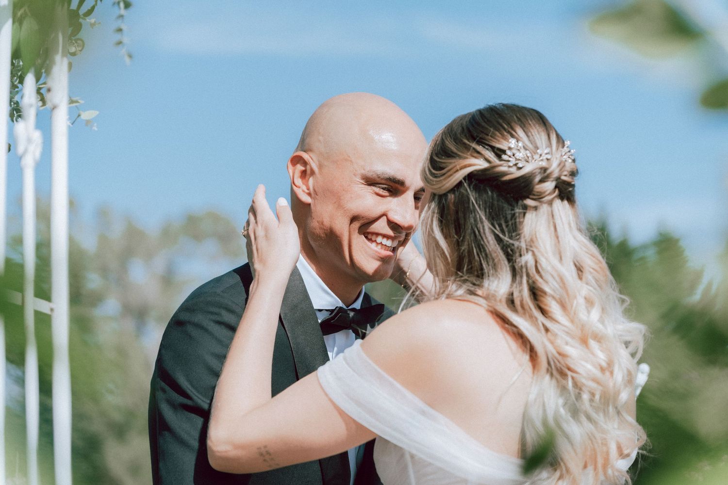 Bald groom smiling joyfully as bride holds his face during outdoor wedding ceremony.