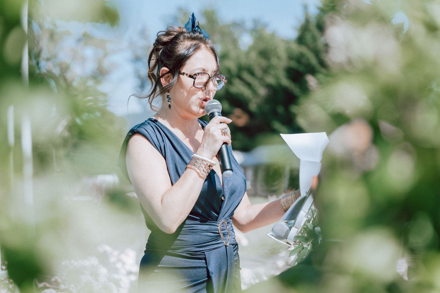 Woman in blue dress speaking into microphone while holding paper during outdoor wedding ceremony.