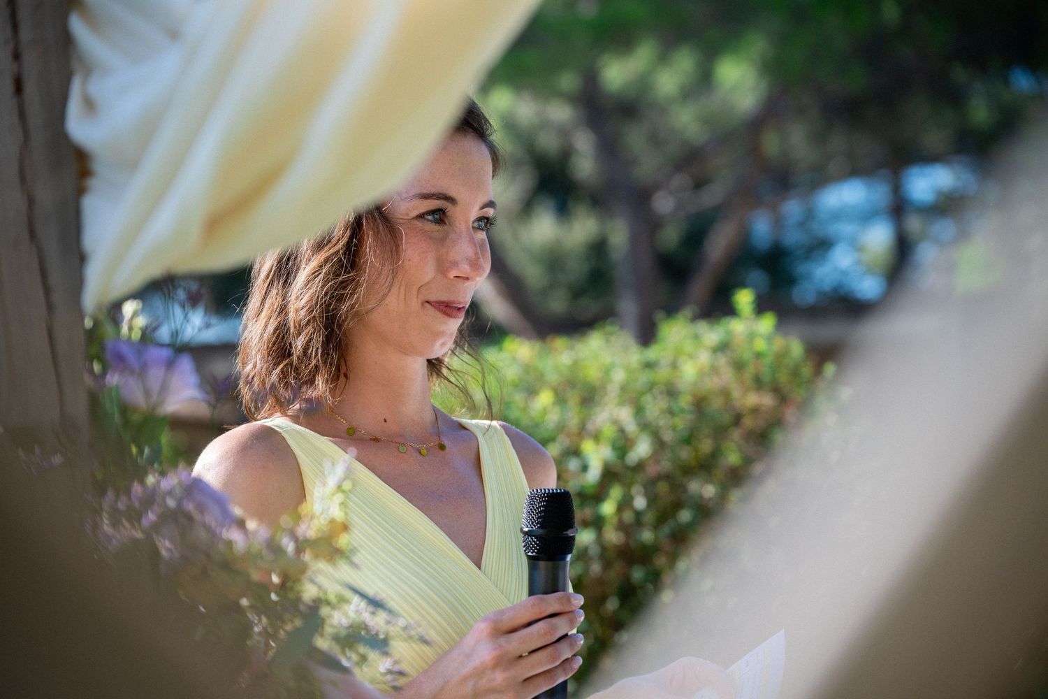 Woman in yellow dress holding microphone speaking outdoors at a garden event.