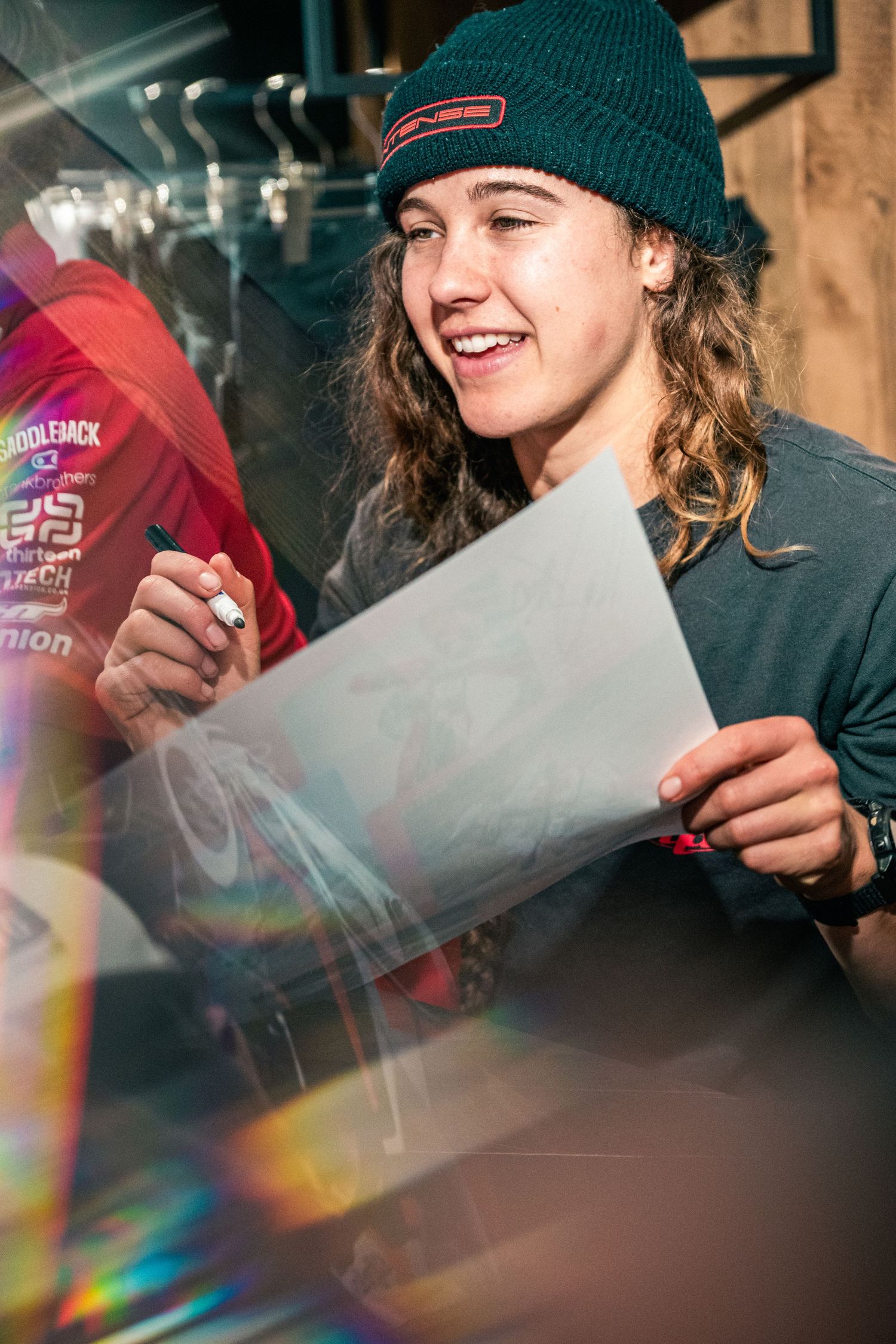 Smiling young person with curly hair and teal beanie signing autograph on paper at a sporting event.