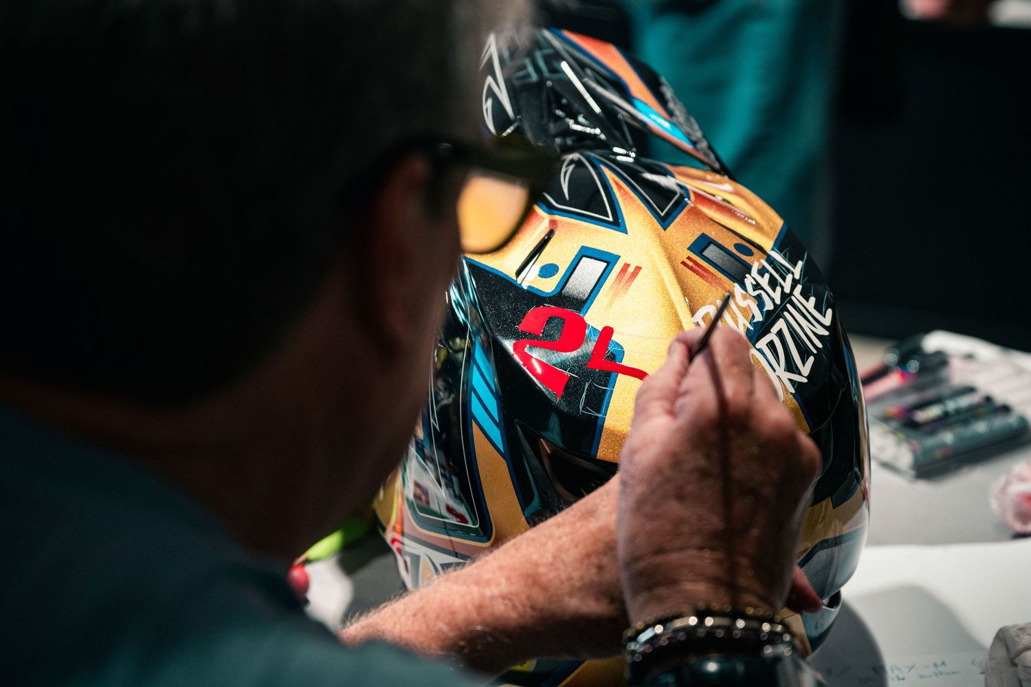 A person autographing a colorful racing helmet with markers in a dimly lit environment.