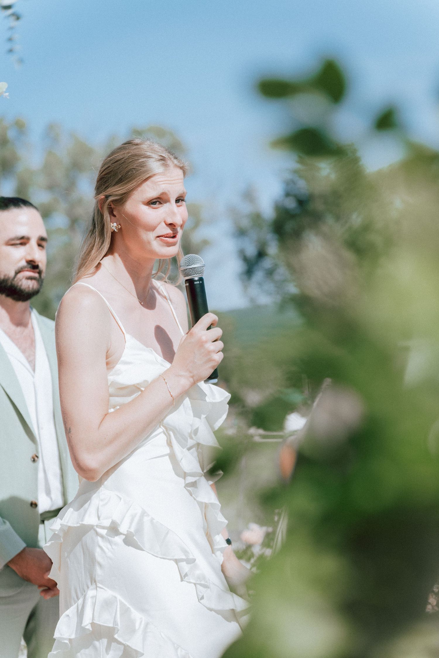 Bride in white ruffled gown speaking into microphone during outdoor wedding ceremony vows.