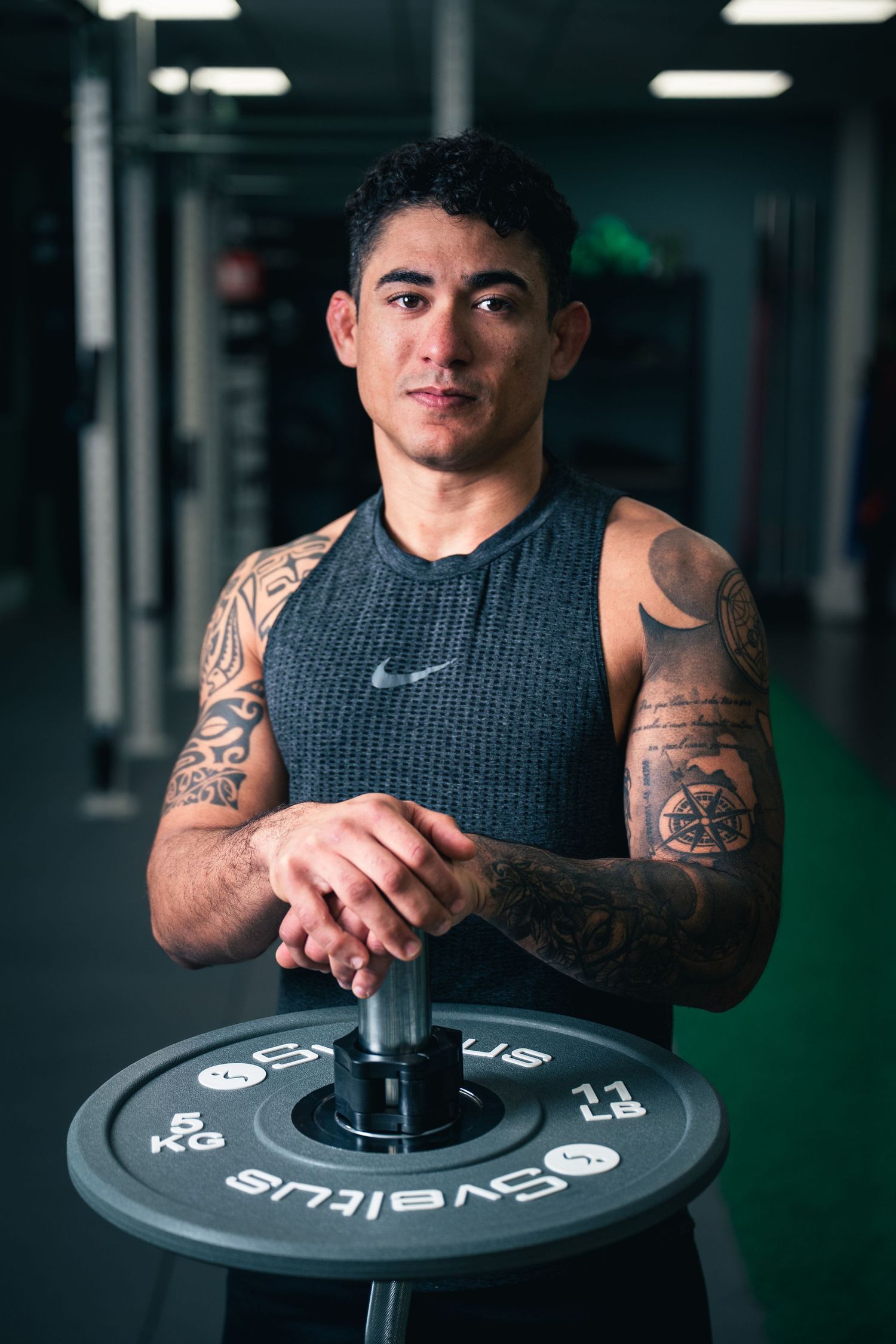 Fit tattooed man in Nike tank top posing with a Sveltus barbell weight plate in gym.