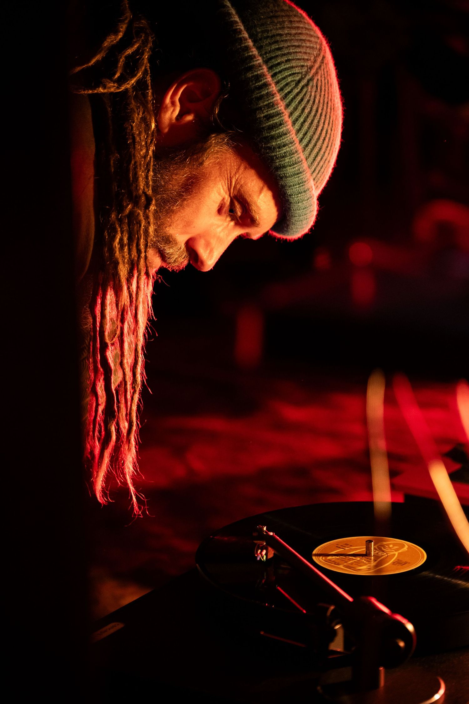 DJ with dreadlocks and beanie hat focused on vinyl record on turntable in red-lit venue.
