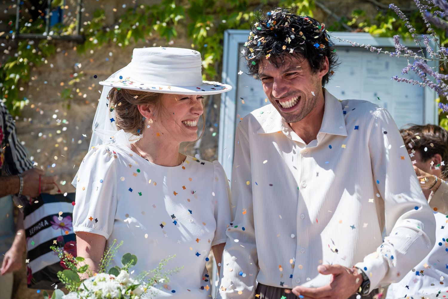 Happy couple laughing together at outdoor wedding celebration with colorful confetti falling around them.