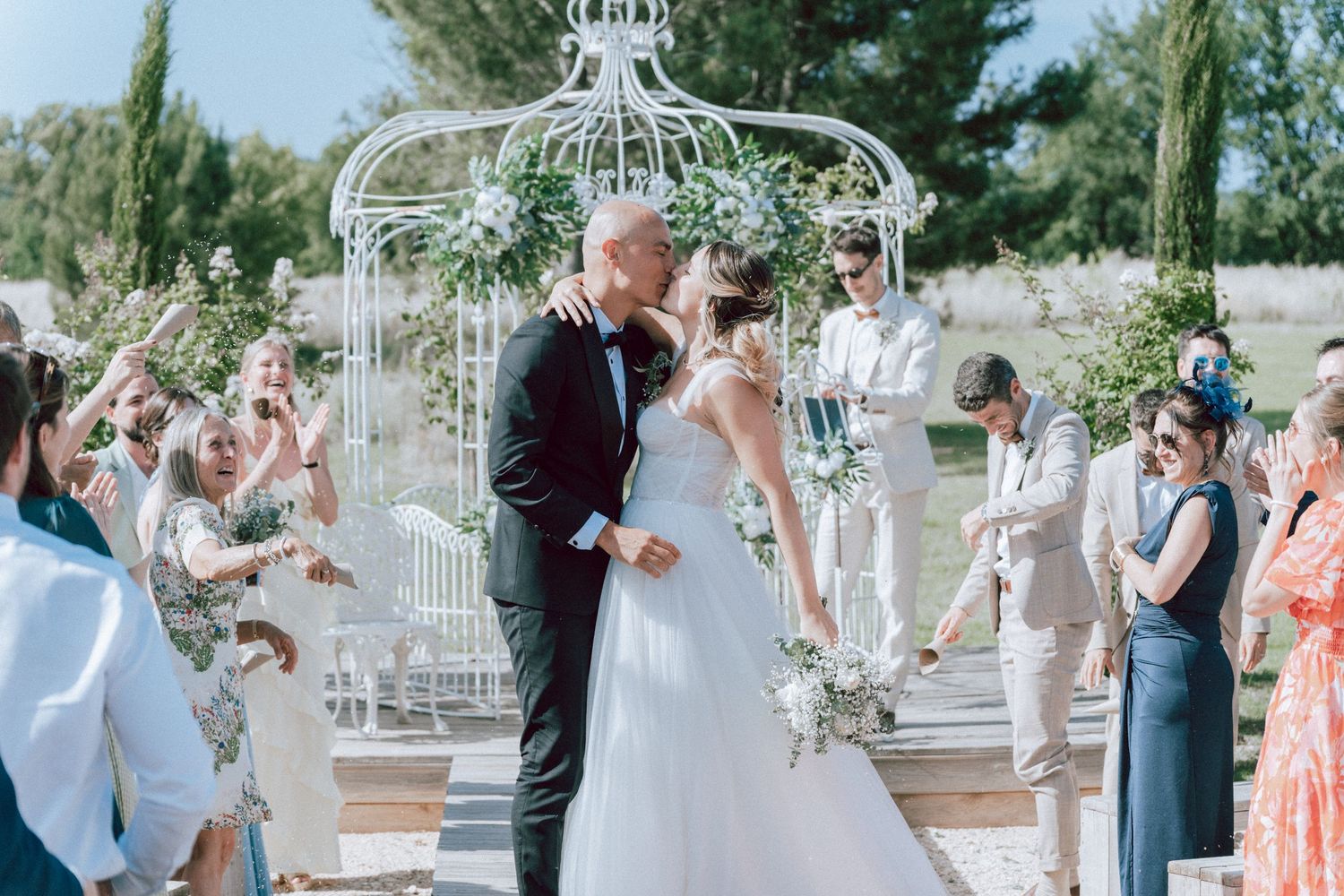 Bride and groom sharing first kiss at altar as guests celebrate outdoor wedding ceremony.