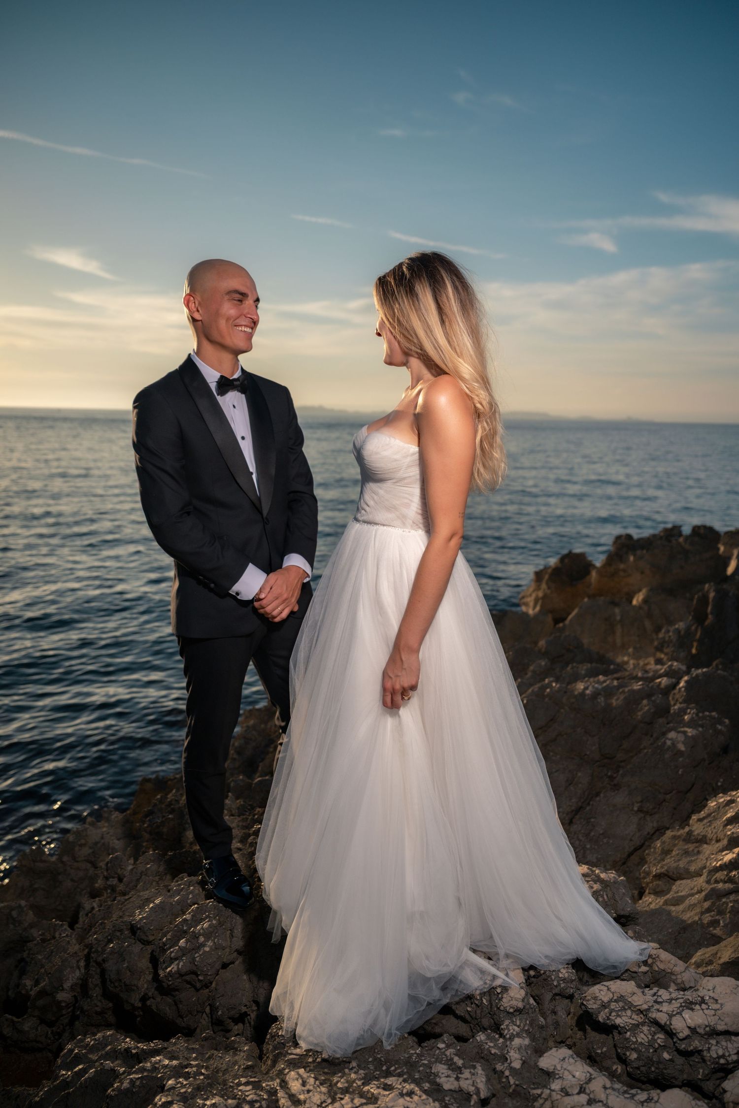 Bride and groom in formal wedding attire stand on rocky coastal cliffs at sunset with ocean backdrop.