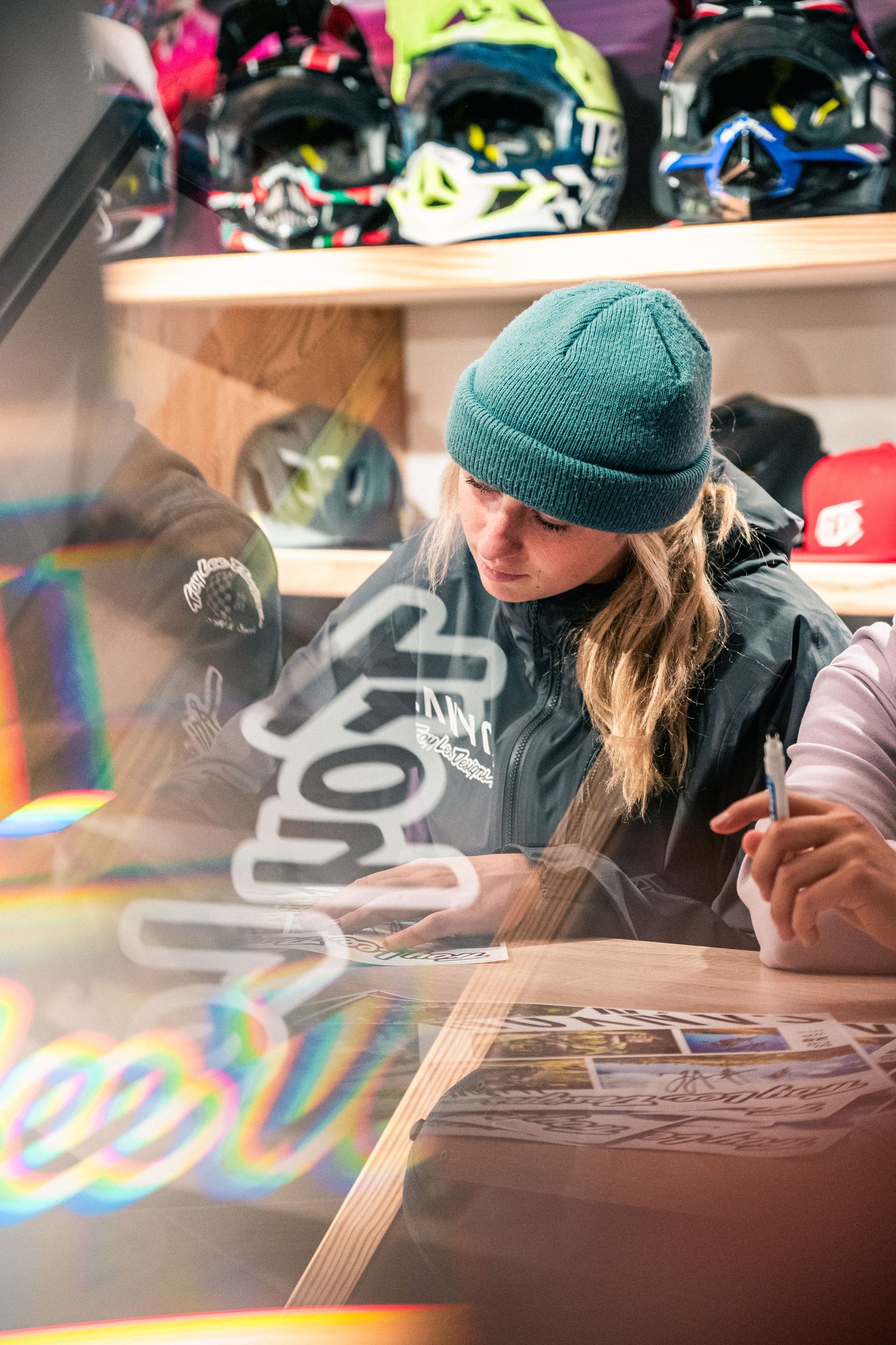 Woman in green beanie signing autographs at a sports shop with colorful helmets displayed on shelves behind her.