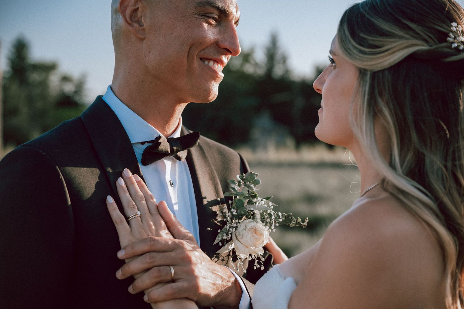 Smiling bald groom gazes at blonde bride while she shows off her wedding ring.