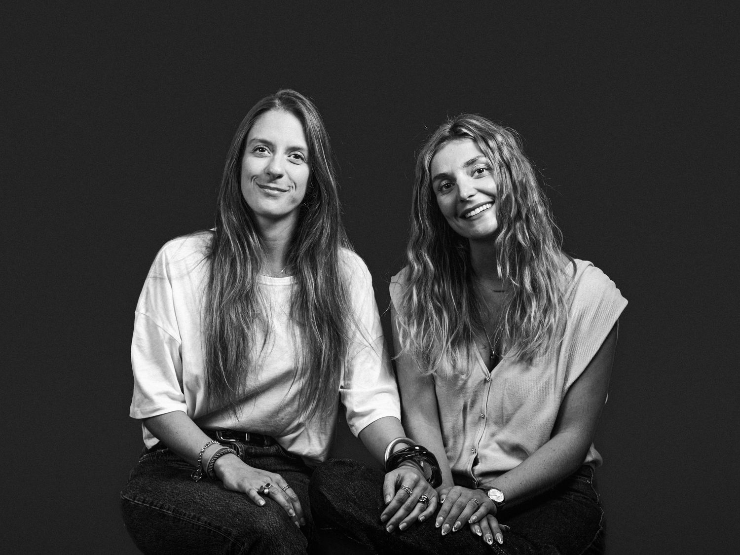 Black and white studio portrait of two young women with long hair sitting together and smiling at the camera.