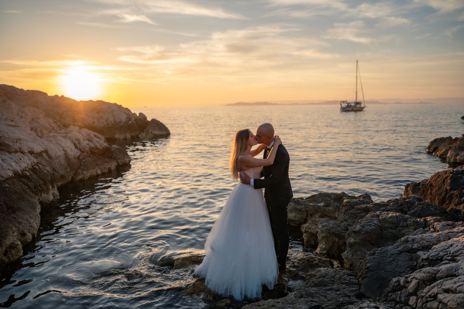 Bride and groom kissing on rocky shore at sunset with sailboat on calm sea in background.