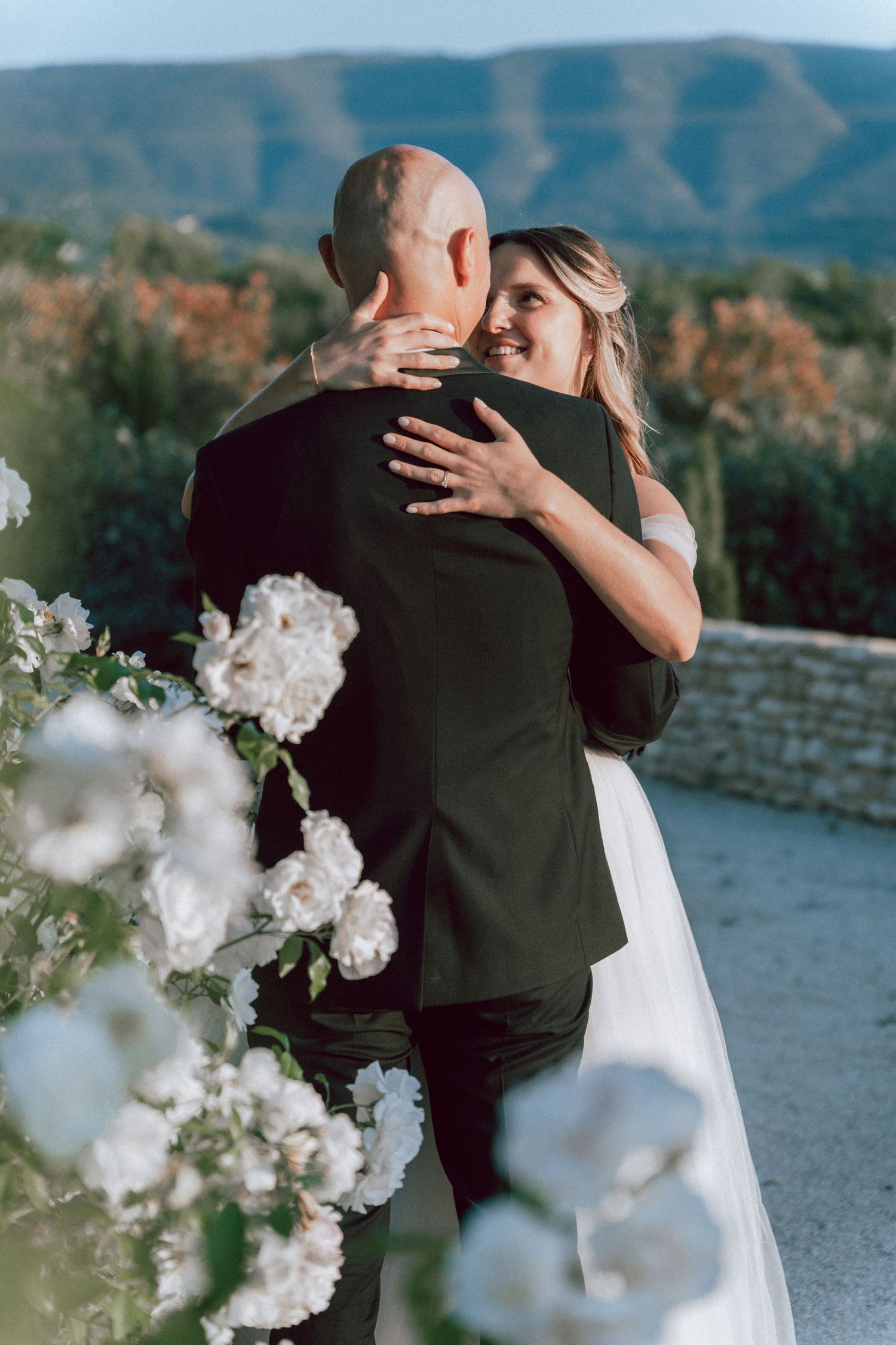 Bride smiles joyfully while embracing groom with mountain landscape and white flowers in background.