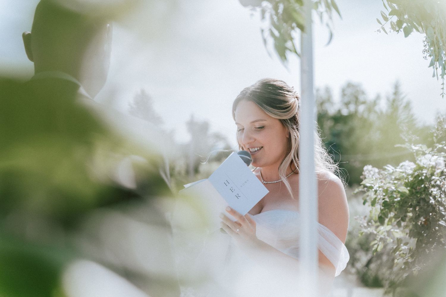 Bride smiling softly while reading vows from card during romantic outdoor wedding ceremony.