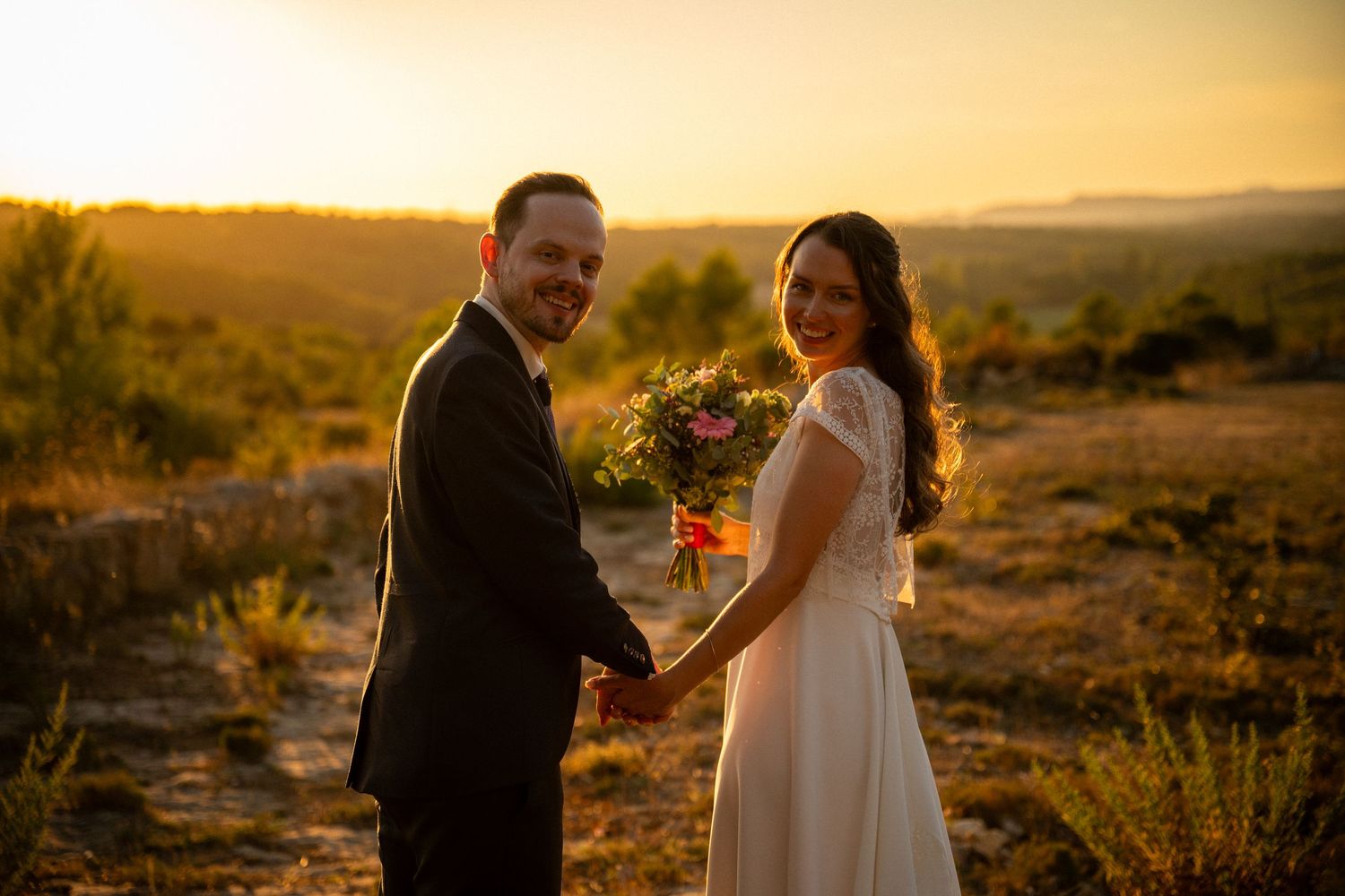 Smiling bride and groom holding hands at sunset in scenic outdoor landscape, bride carrying pink bouquet.
