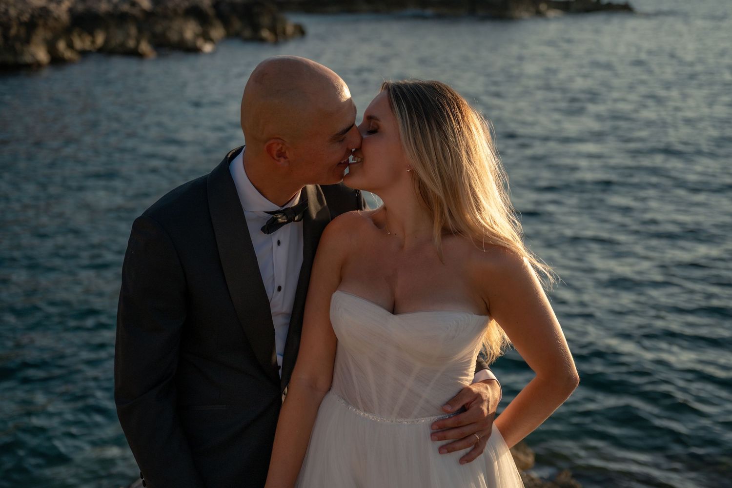 A bride and groom share a romantic kiss by the ocean at sunset, dressed in wedding attire.