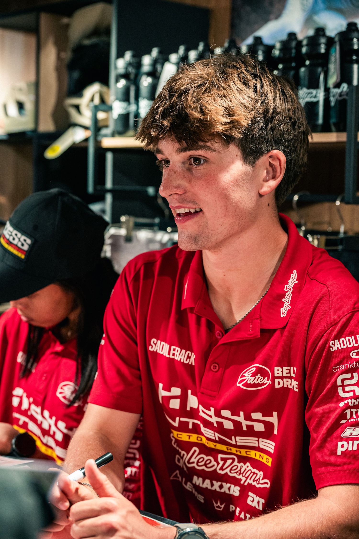 Young man in red Saddleback racing team polo shirt smiling at motorsport signing event.