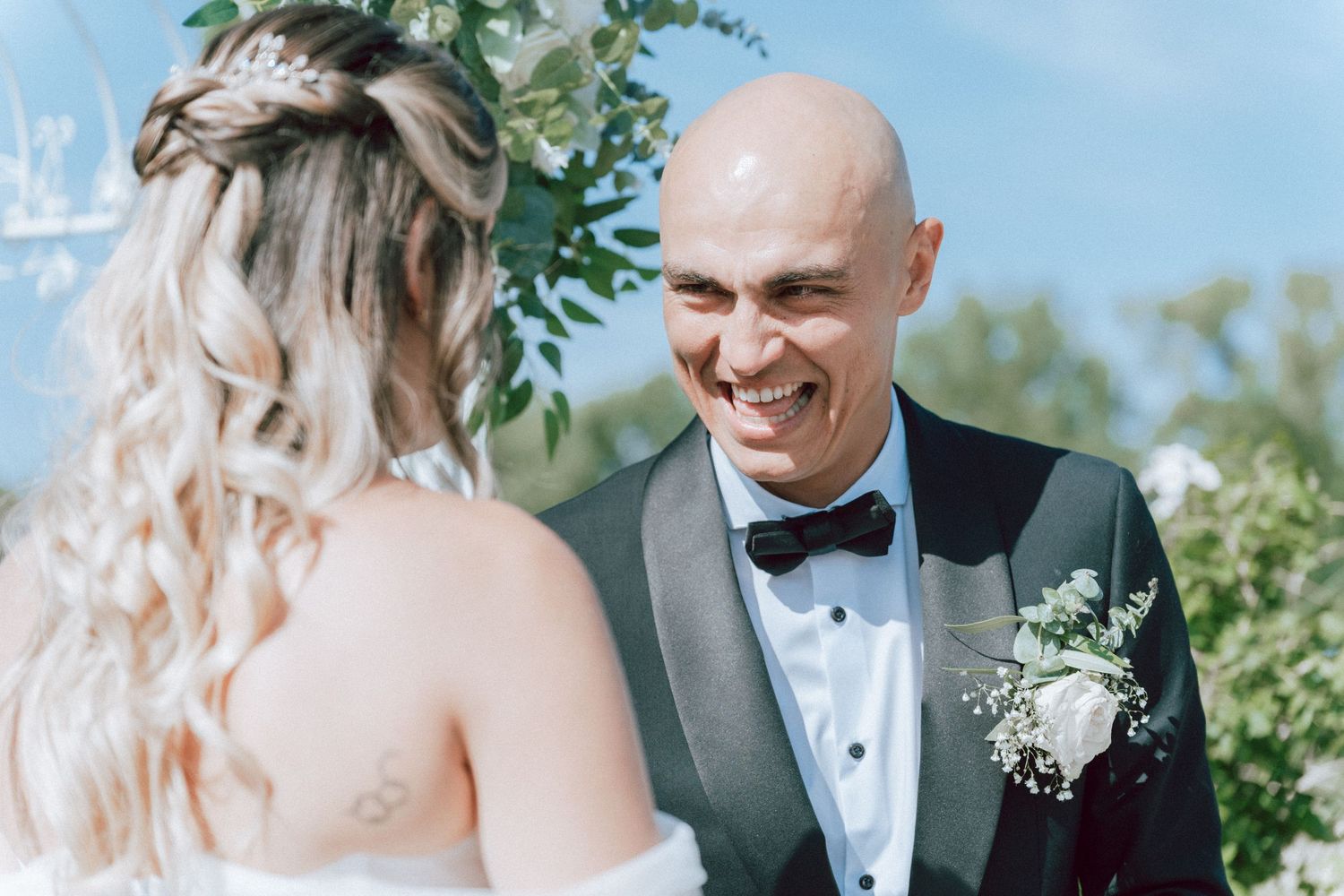 Smiling bald groom in black tuxedo with boutonniere faces bride at outdoor wedding ceremony.
