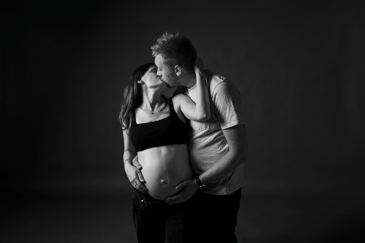 Black and white maternity photo of a couple sharing a tender kiss while cradling a pregnant belly in a studio.
