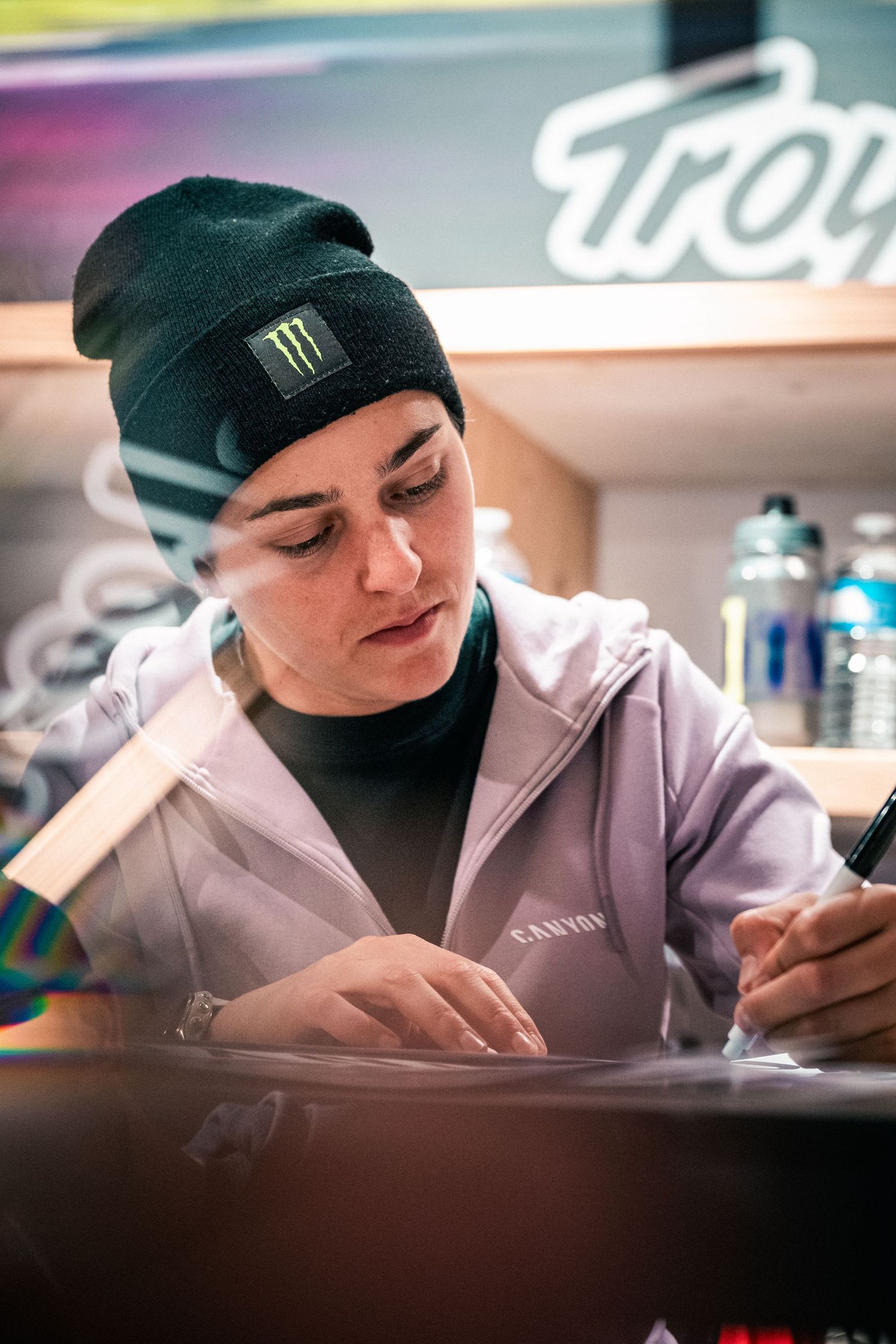 Young woman in Monster Energy beanie and Canyon hoodie signing autographs at a motorsport event.