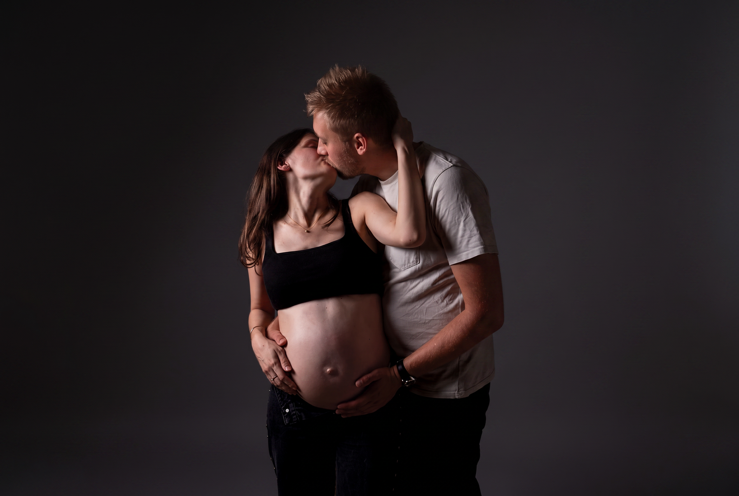 Couple sharing a tender kiss during a maternity photo shoot, showcasing a pregnant belly against a dark studio background.