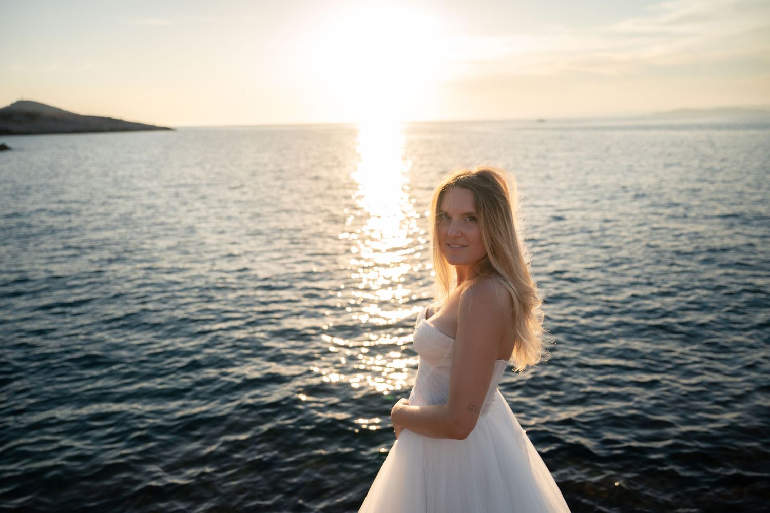 Beautiful bride in white gown smiling by the ocean during a golden sunset.