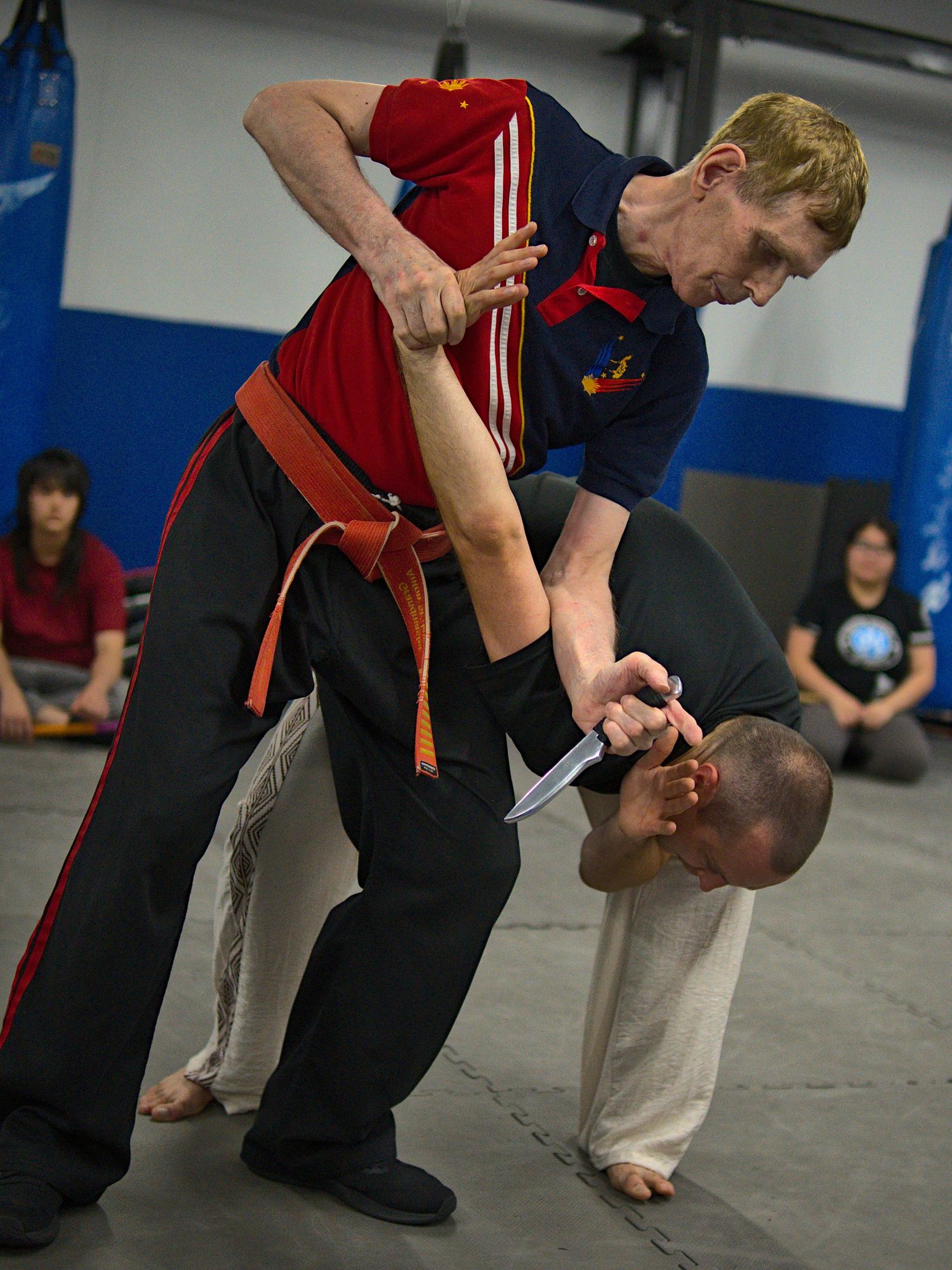 Martial arts instructor demonstrates knife defense technique on kneeling student in dojo.
