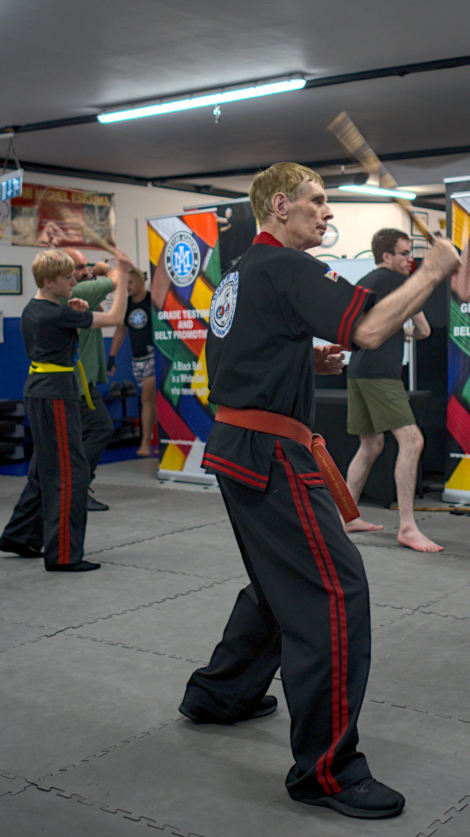 Martial arts instructor in black uniform leads students in weapon training drill at indoor dojo.