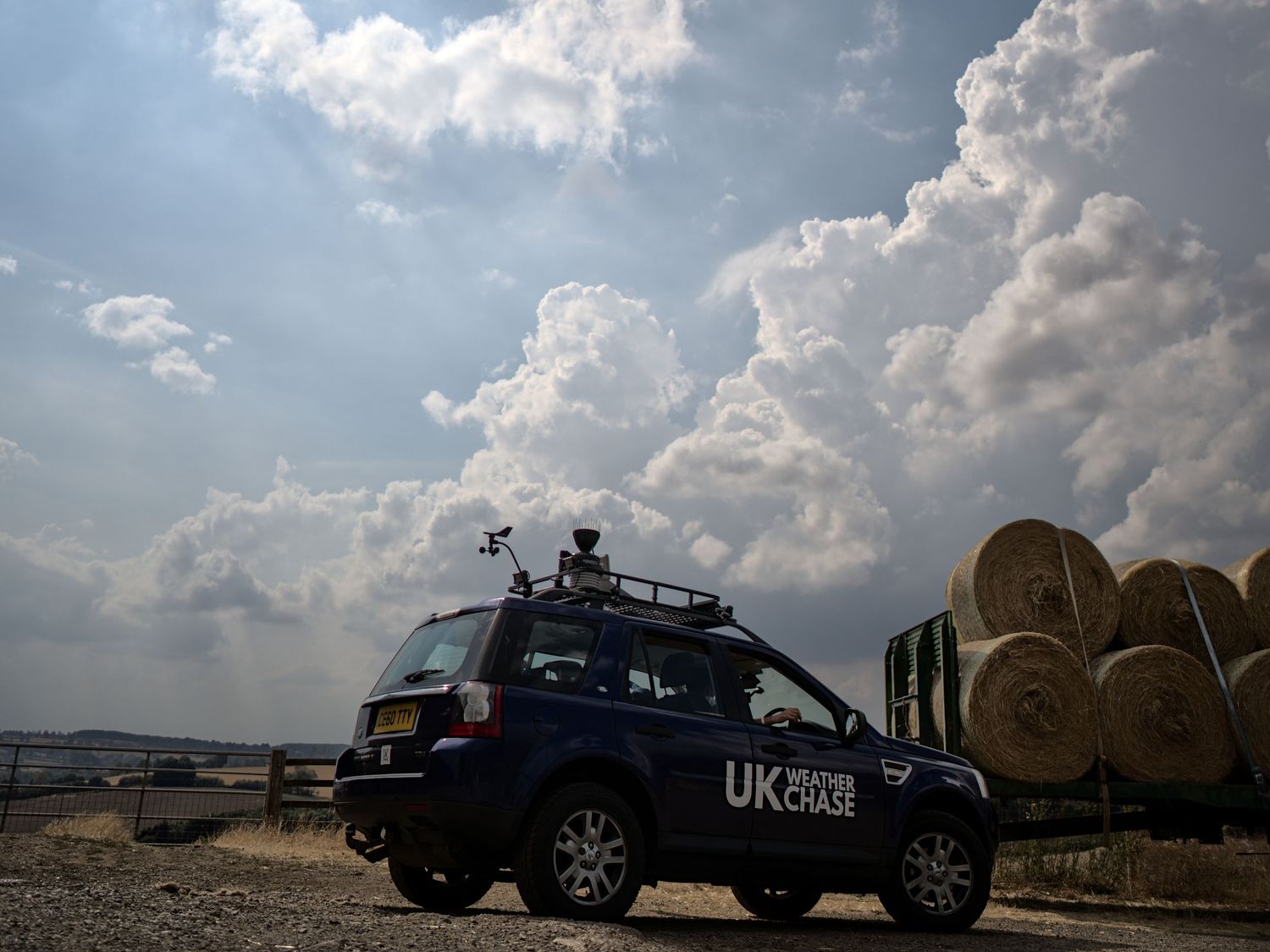 UK Weather Chase Land Rover parked near hay bales on a farm under dramatic storm clouds in England.
