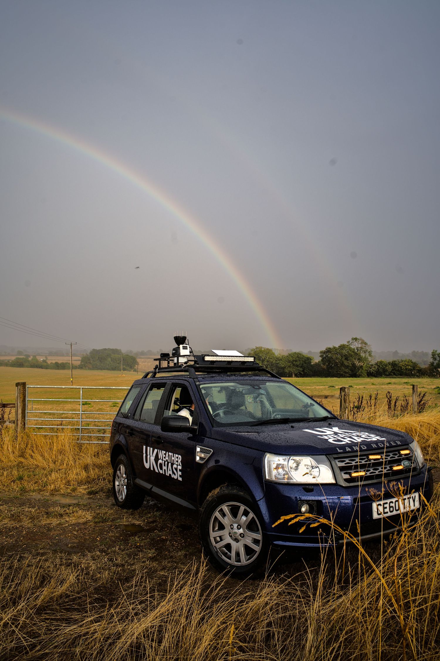 Blue Land Rover SUV with rooftop equipment parked in a field under a vibrant rainbow in a cloudy sky.