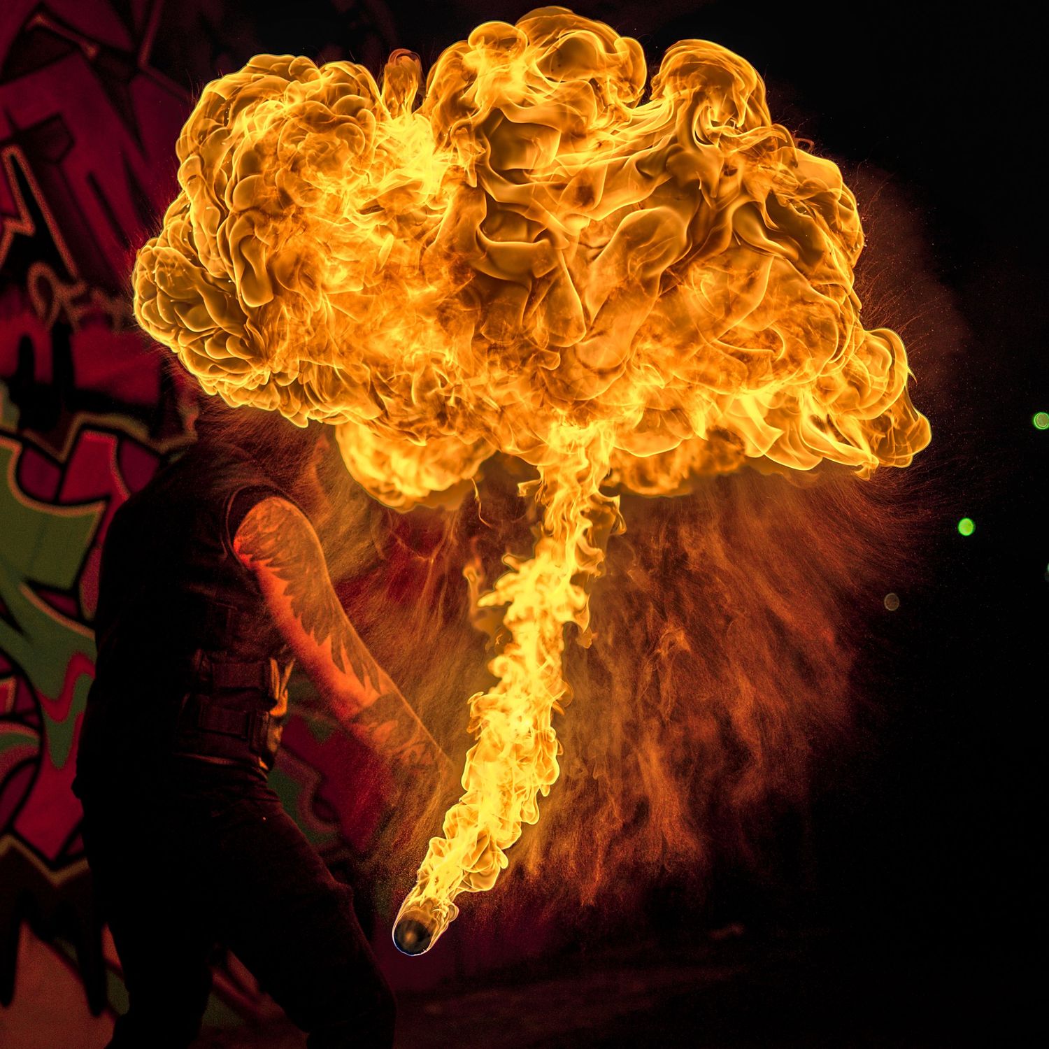 Street performer breathes fire creating large mushroom cloud flame against colorful graffiti wall.