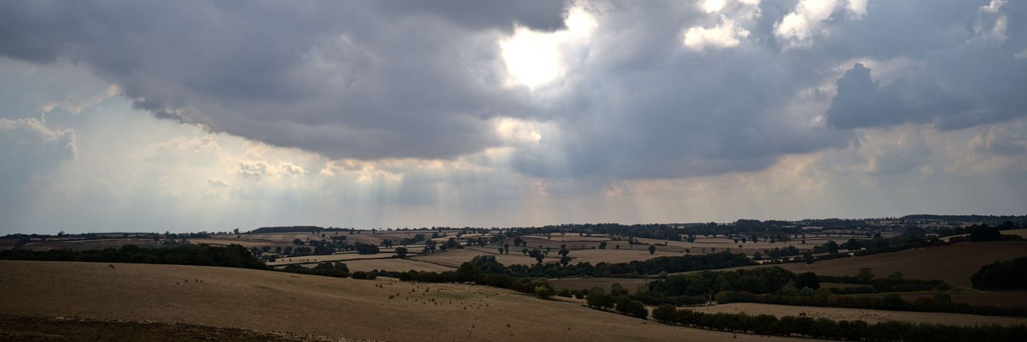 Panoramic view of rolling English countryside with fields and villages beneath dramatic overcast storm clouds.