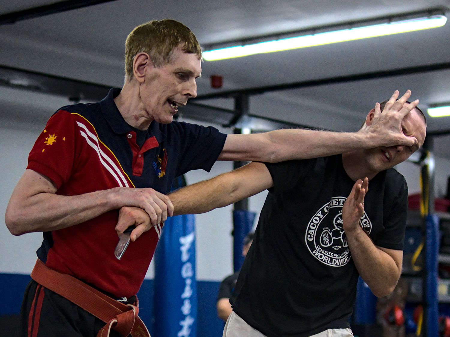 Two martial artists practice knife defense techniques together in a professional indoor training facility.