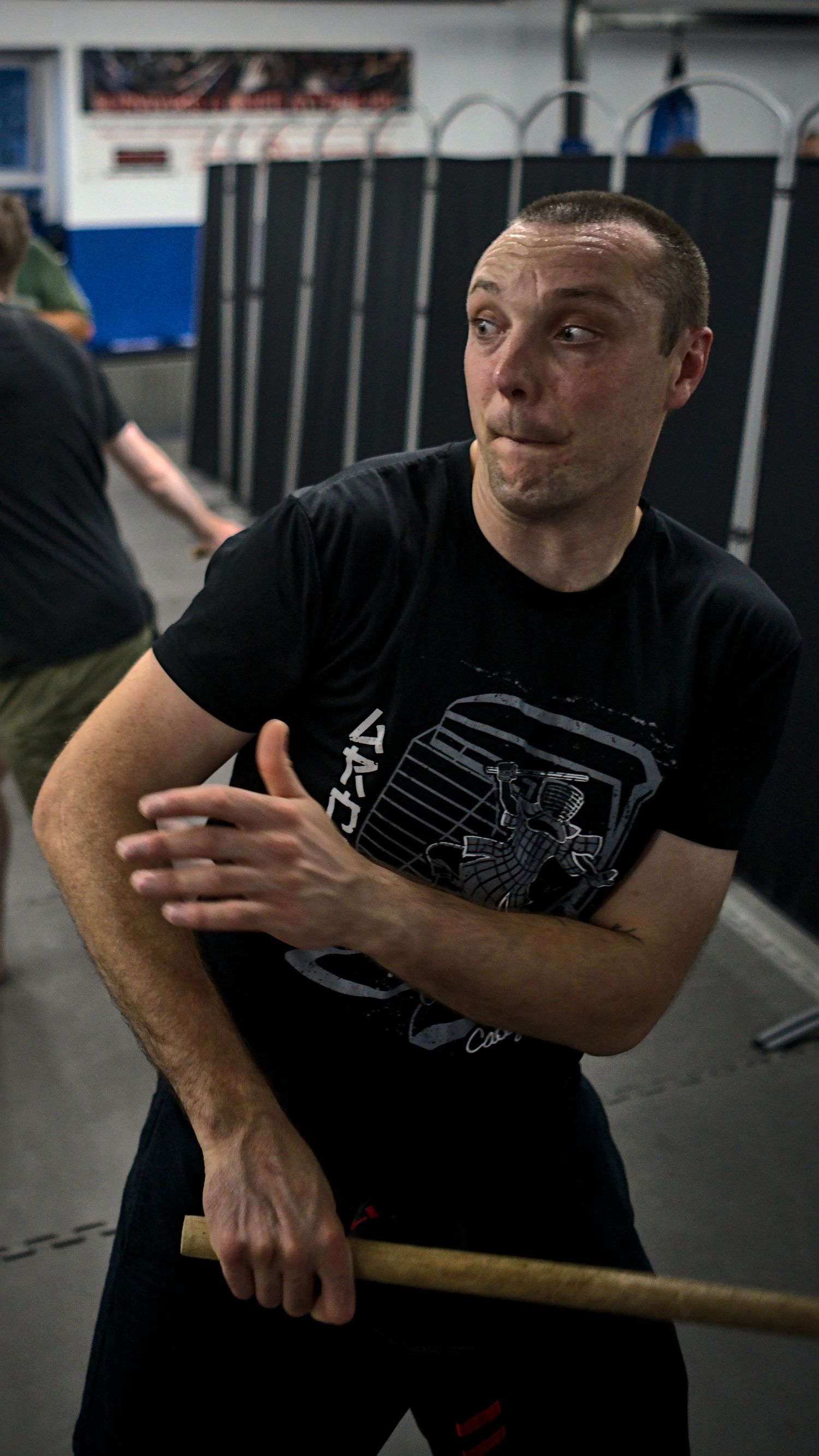 A man in a black t-shirt practices stick fighting defensive techniques in a martial arts gym.