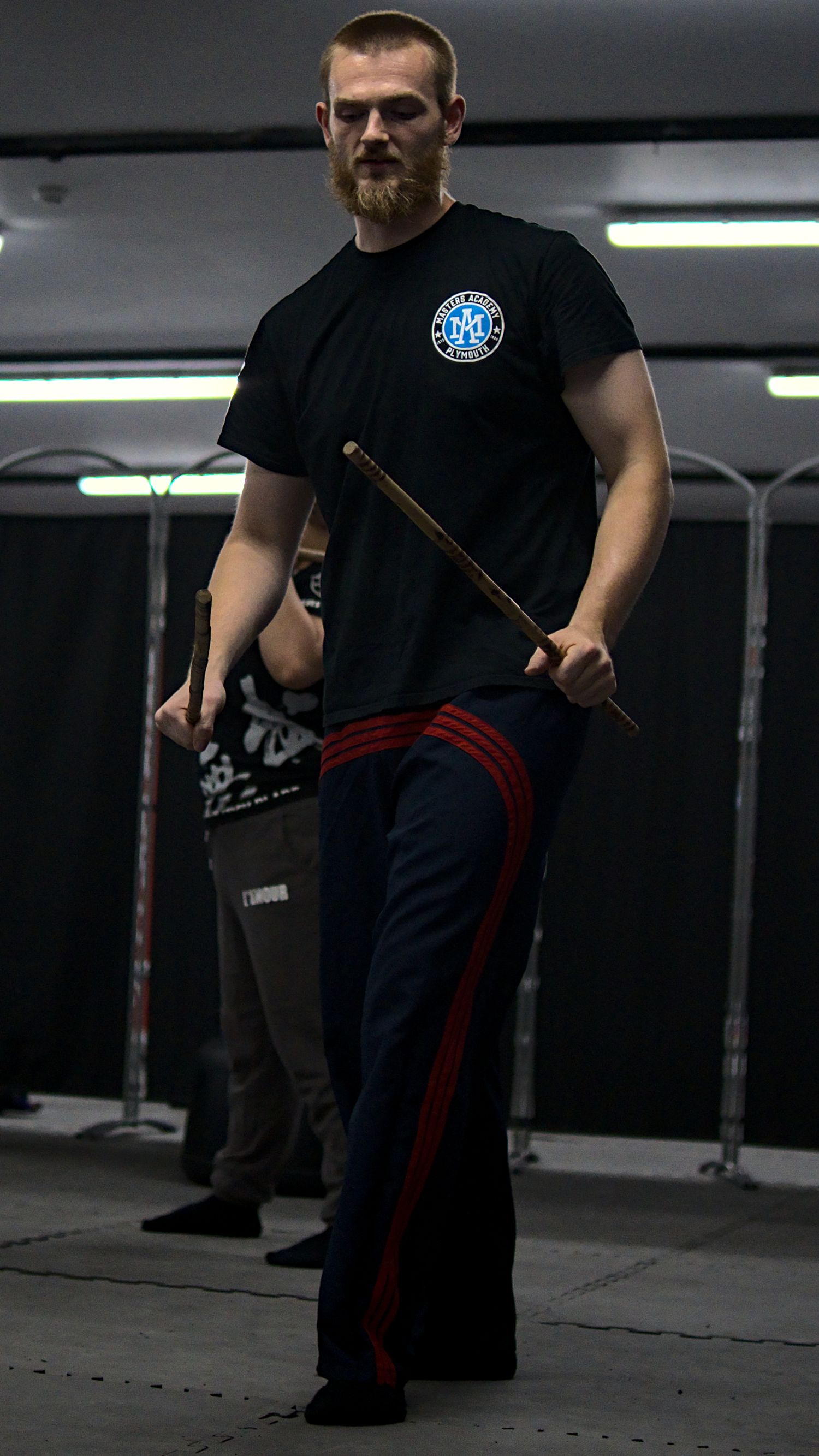 A bearded man in a black martial arts shirt holds a rattan stick during training.