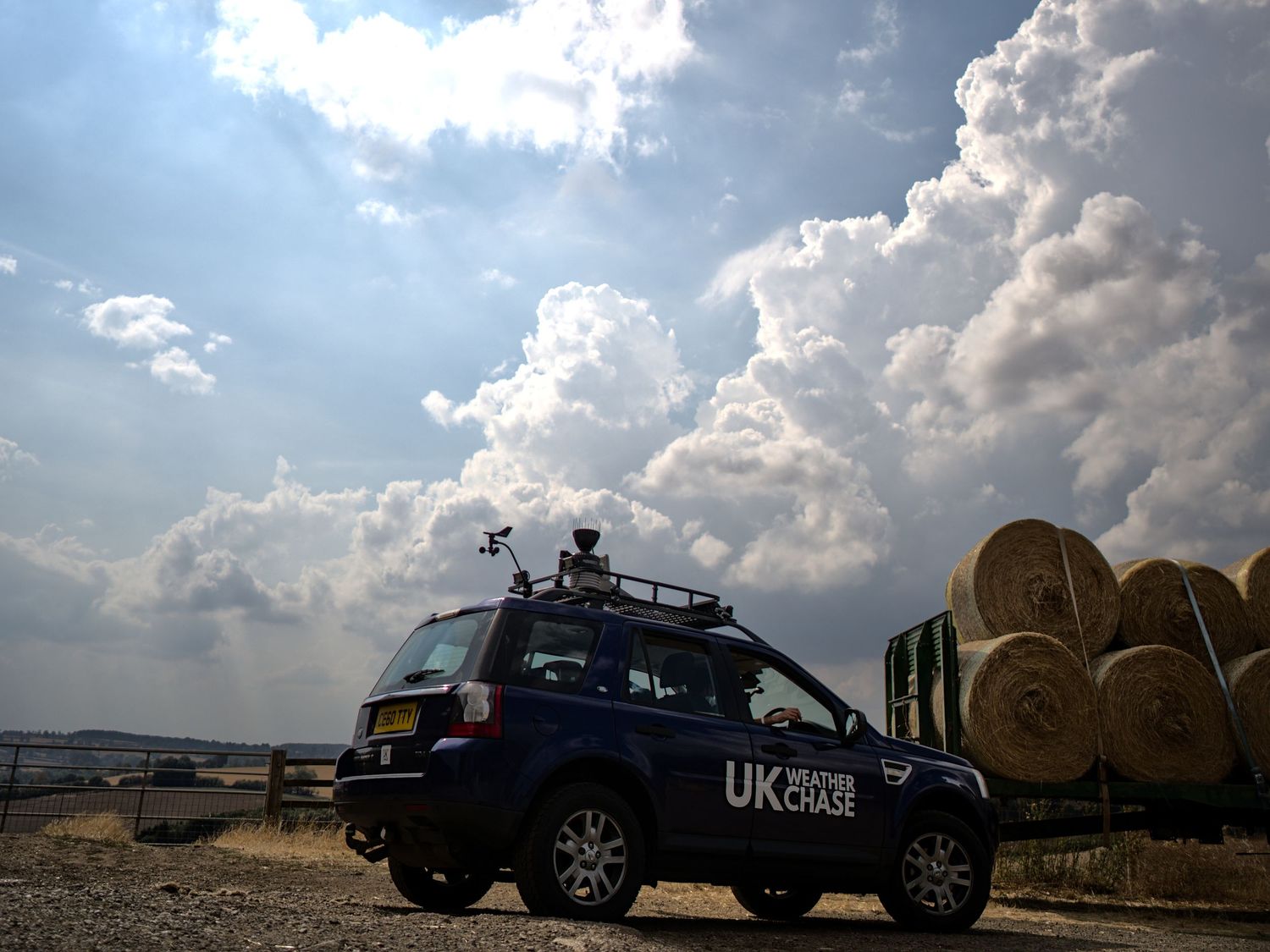 UK Weather Chase Land Rover parked beside large hay bales on a farm under dramatic cumulus clouds.