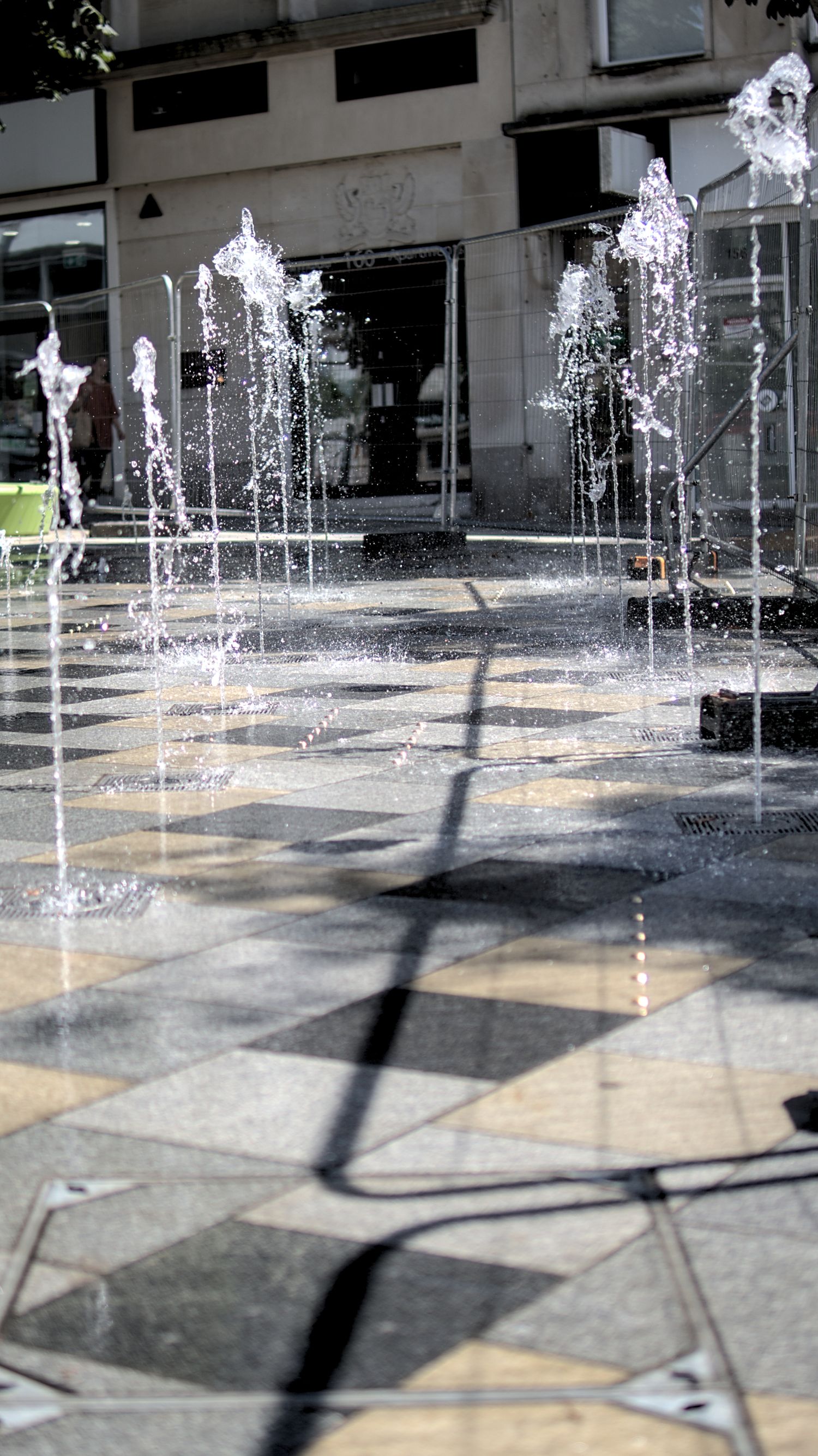 Ground-level fountains shooting water jets on a checkered plaza in bright sunlight.
