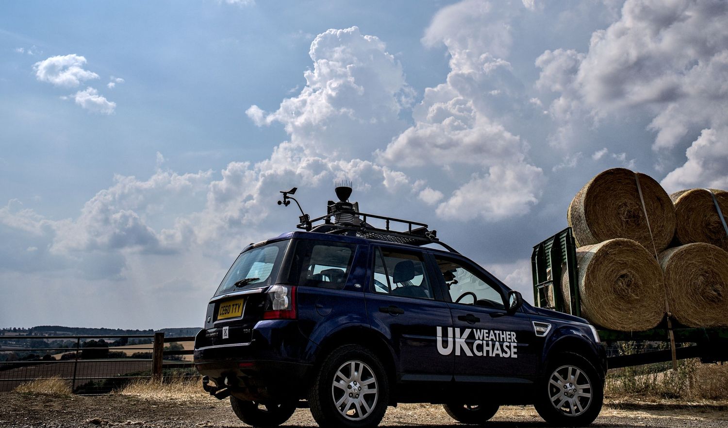 UK Weather Chase Land Rover beside hay bales on a farm under towering cumulus clouds in England.