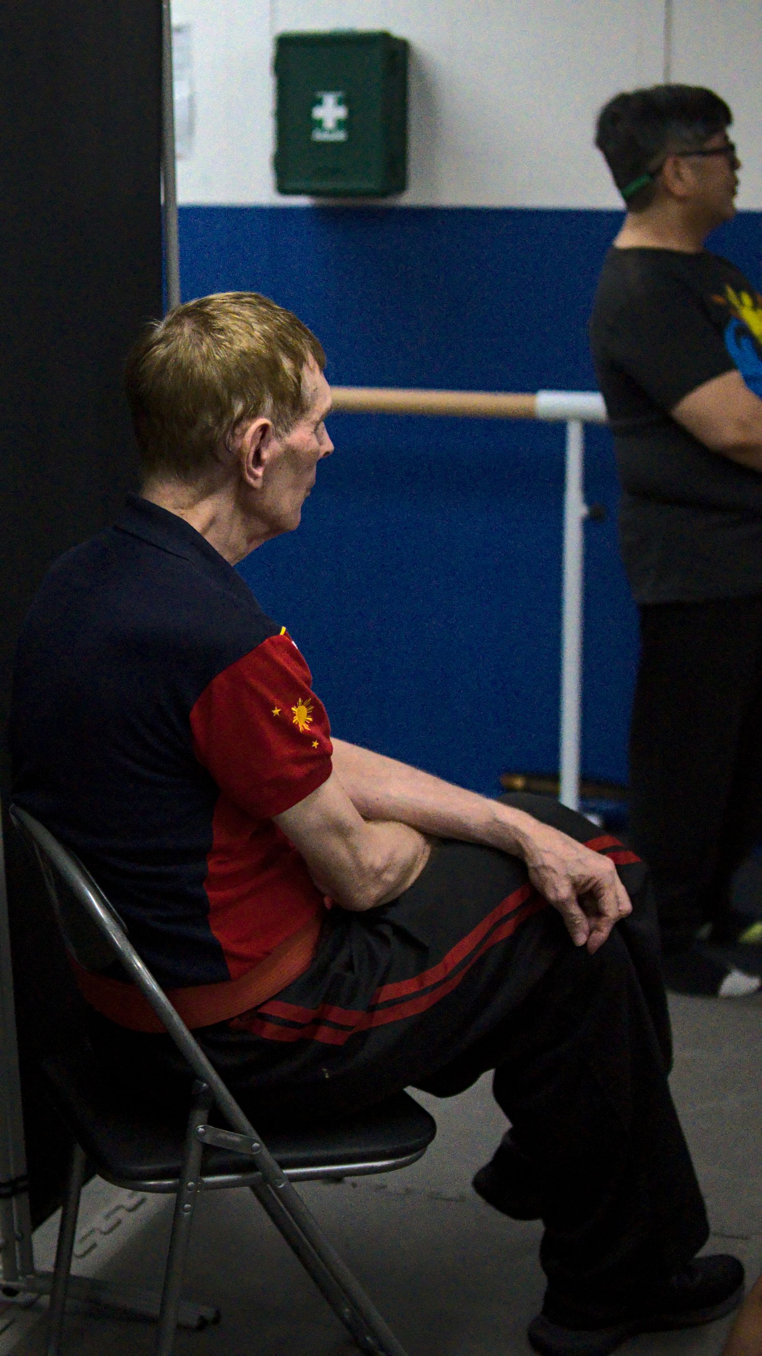 Martial arts instructor in red and black uniform sits observing class at indoor training facility.
